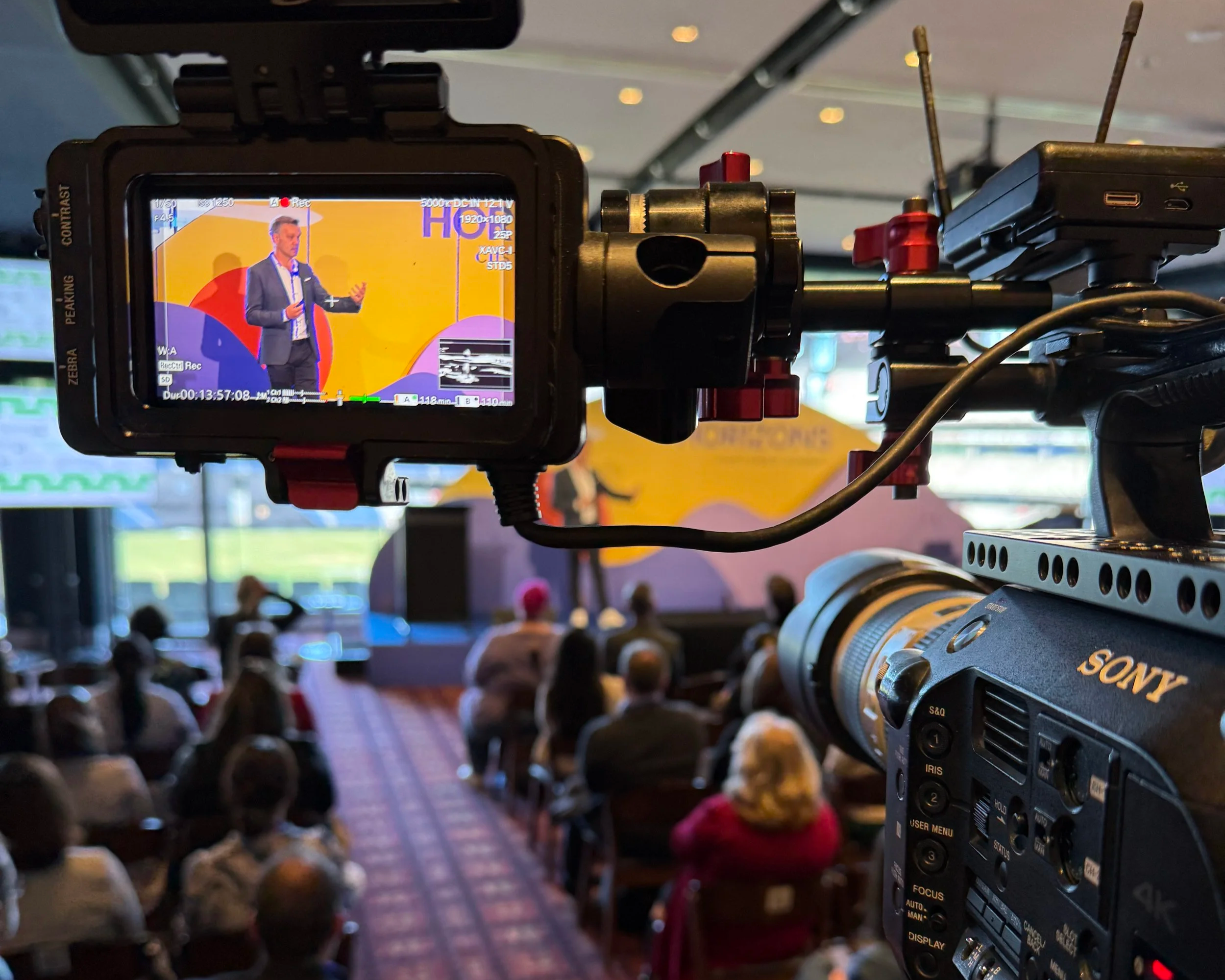 A camera viewfinder screen showing a man in a suit giving a presentation on stage with a yellow and purple background, while the audience is seated and watching.