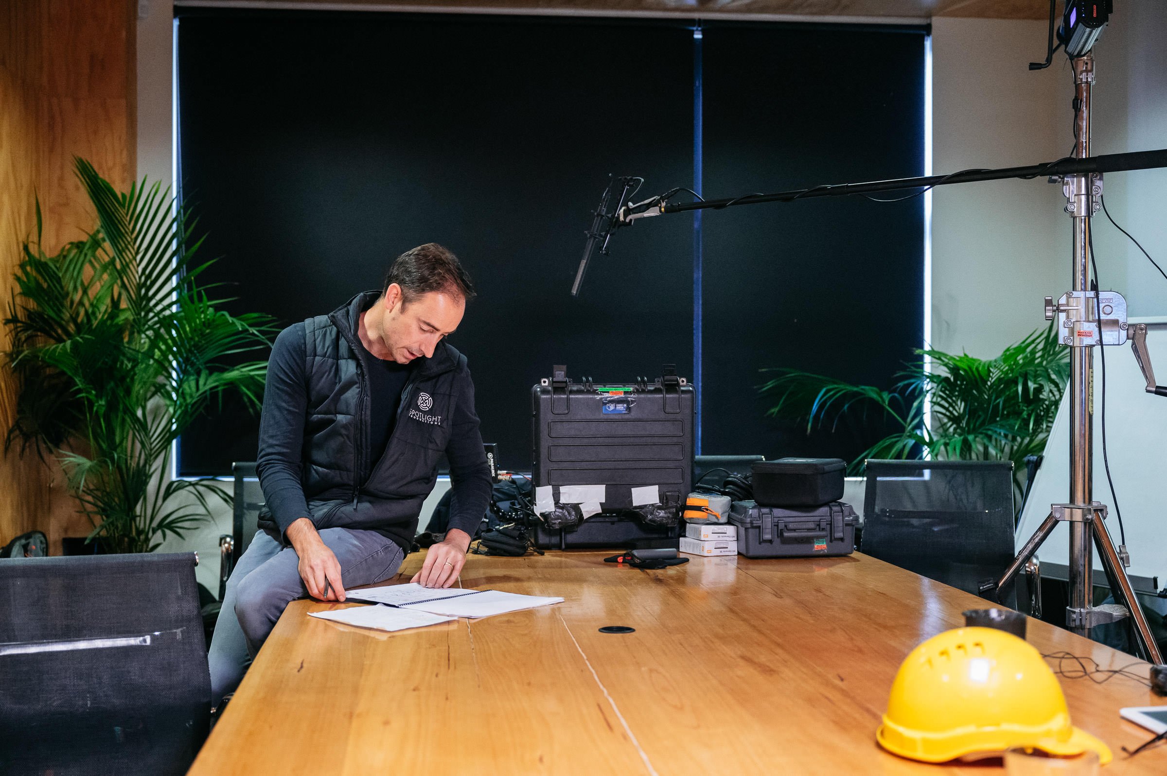 A man in a black vest and gray pants sitting at a wooden table, surrounded by equipment and plants, preparing for a recording or photo shoot in a studio with a black backdrop and professional lighting setup.