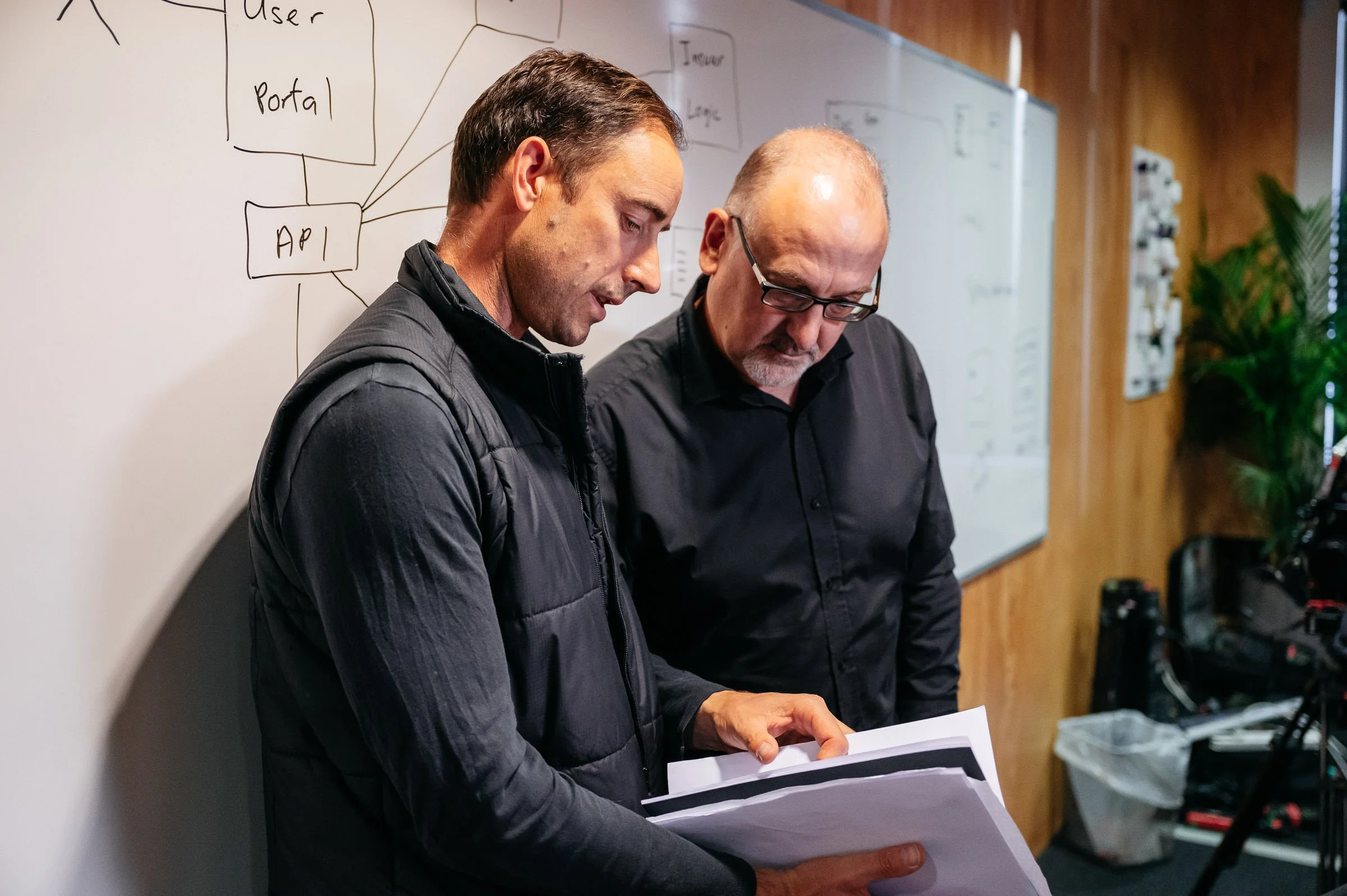 Two men standing near a whiteboard, looking at a document or folder. The whiteboard behind them has a diagram with labeled boxes and lines, including words like 'API', 'Portal', 'User', and 'Login'. The setting appears to be a meeting or discussion room with wooden walls and some plants.