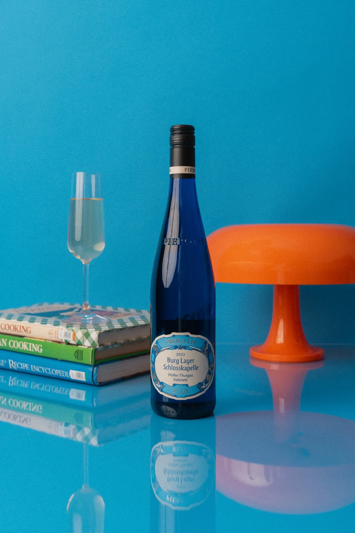 A blue bottle of wine, a glass of wine, a stack of cookbooks, an orange cake stand, all on a reflective blue surface against a blue background.