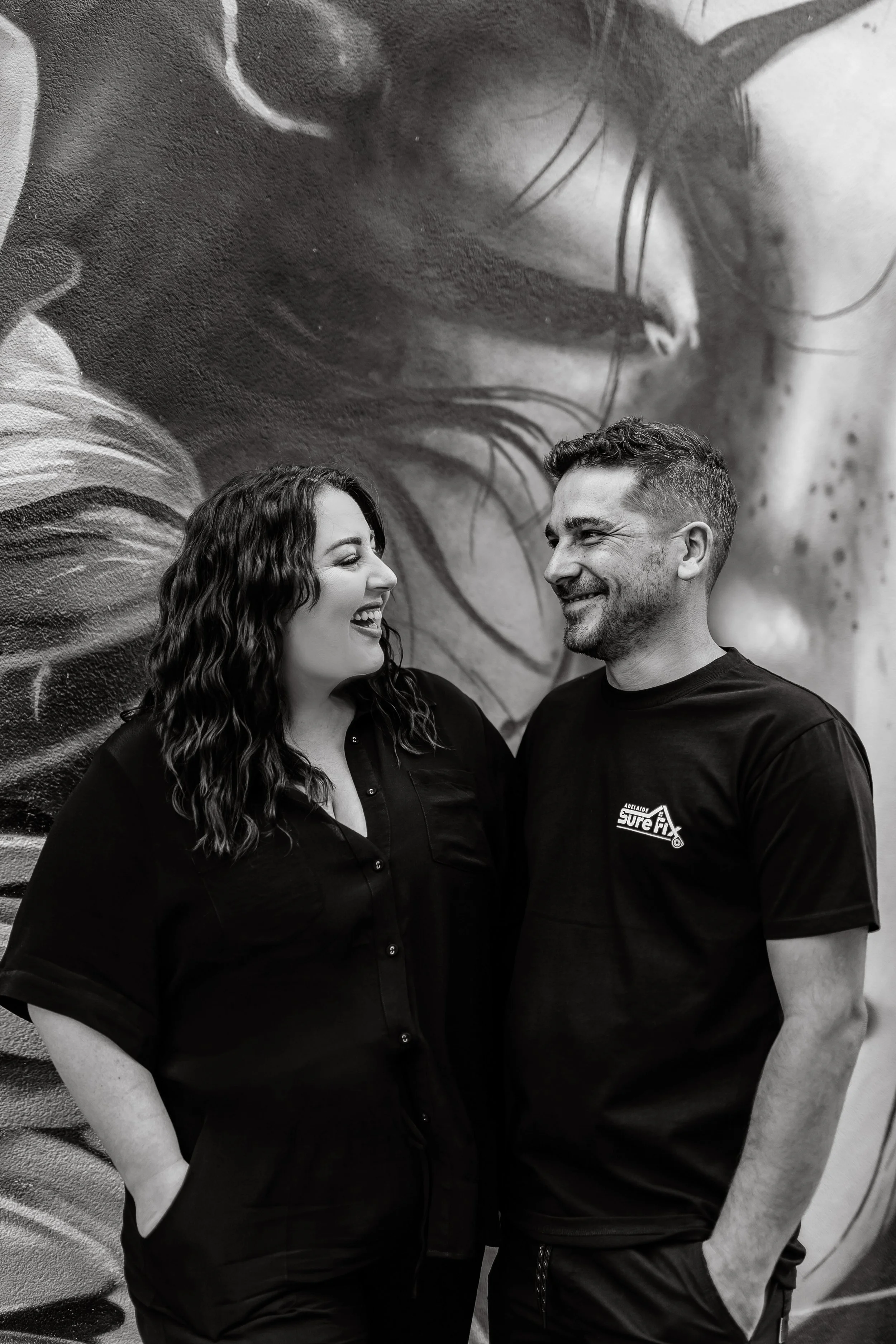 A woman and man smiling and looking at each other, standing in front of a mural of a face with closed eyes.