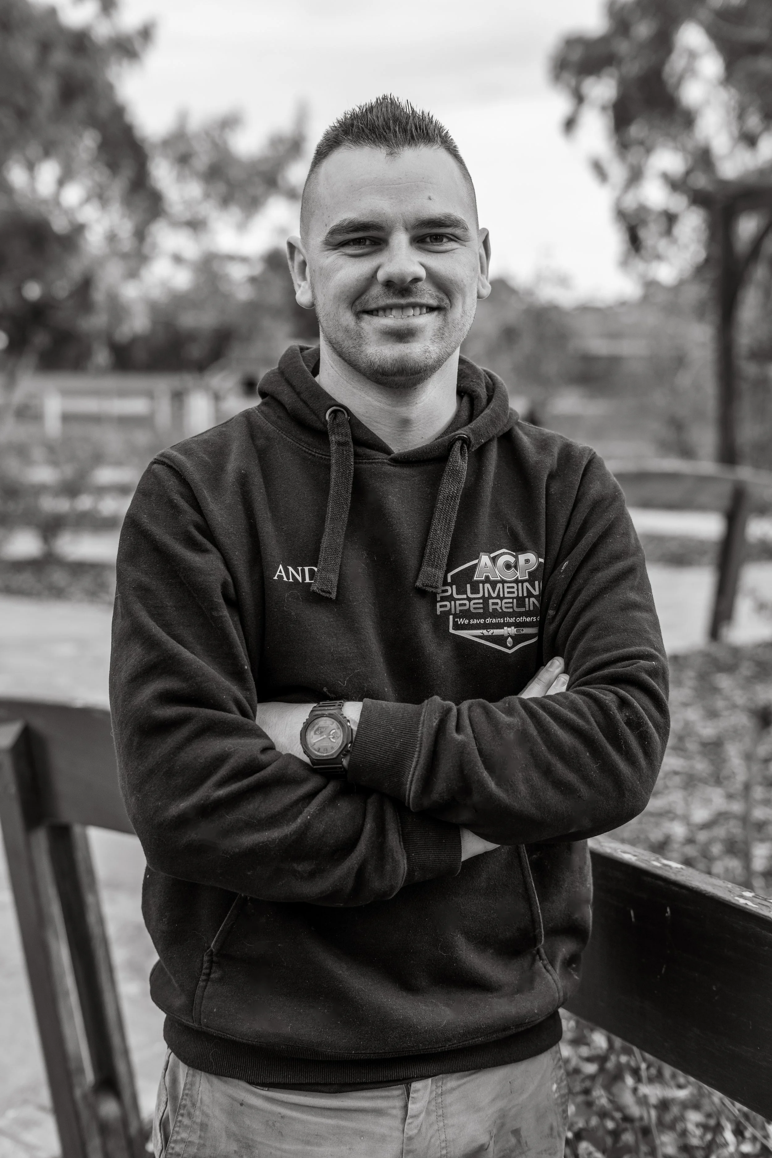 A young man with short hair and a beard stands outdoors with arms crossed, smiling at the camera. He is wearing a dark hoodie and a wristwatch.