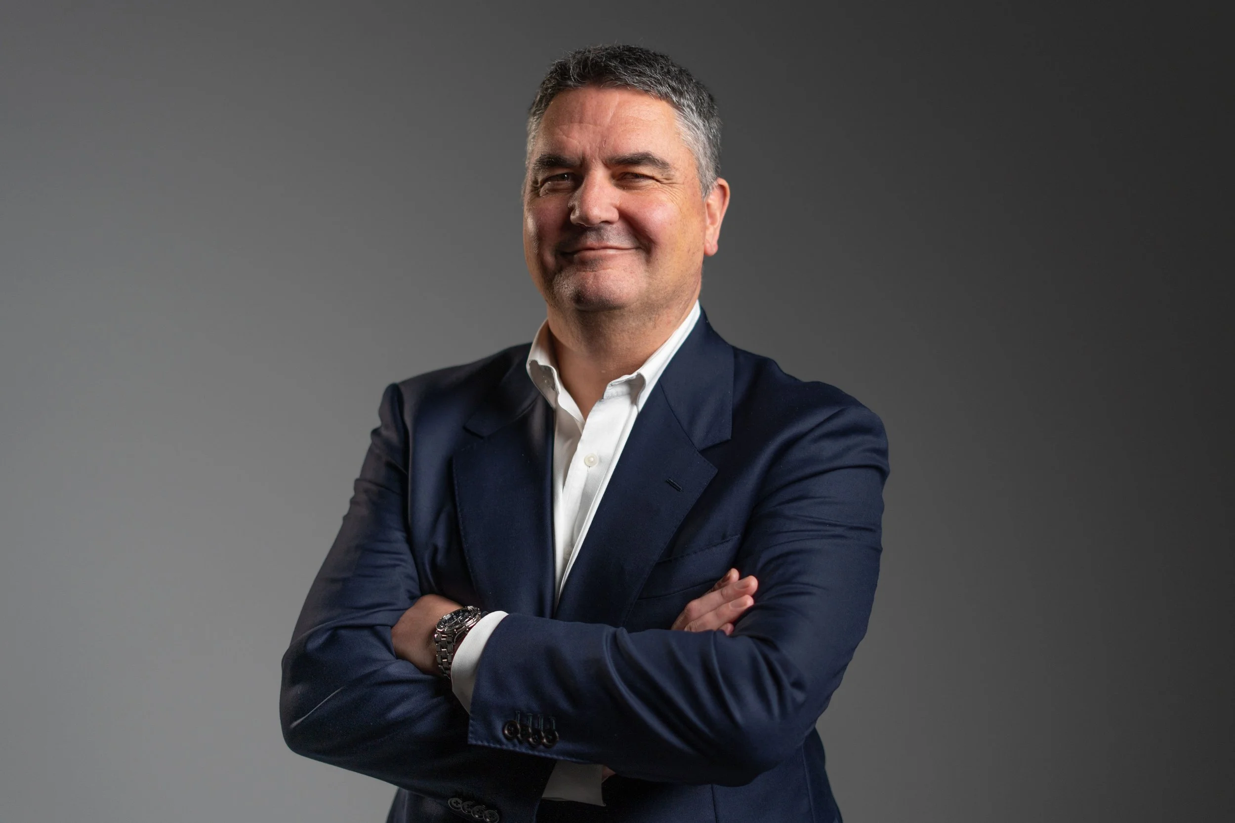 A middle-aged man with short, graying hair, wearing a white shirt and a navy blue blazer, standing with arms crossed and smiling against a gray gradient background.