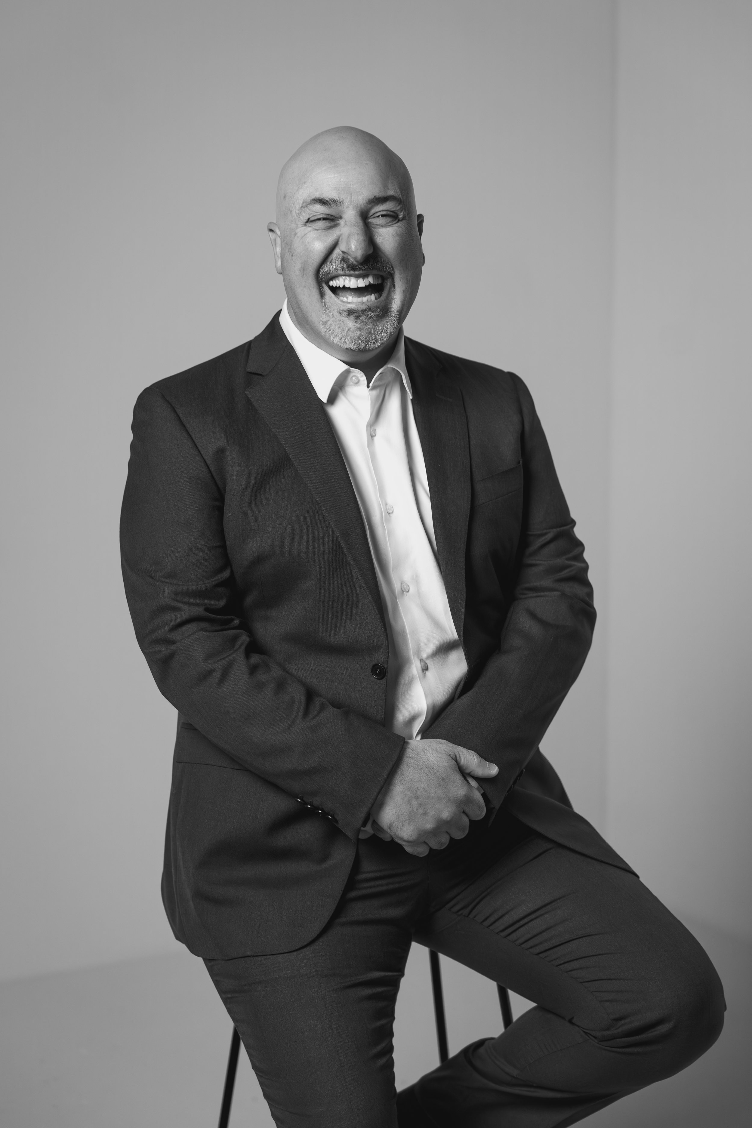 black and white headshot of a man in a suit, sitting on a stool.