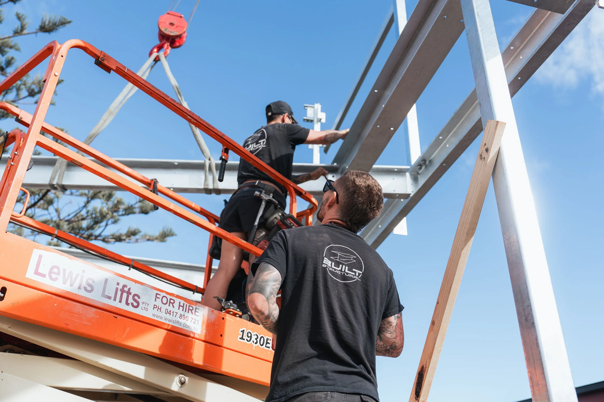 Two workers on an orange lift working on a metal structure outdoors.