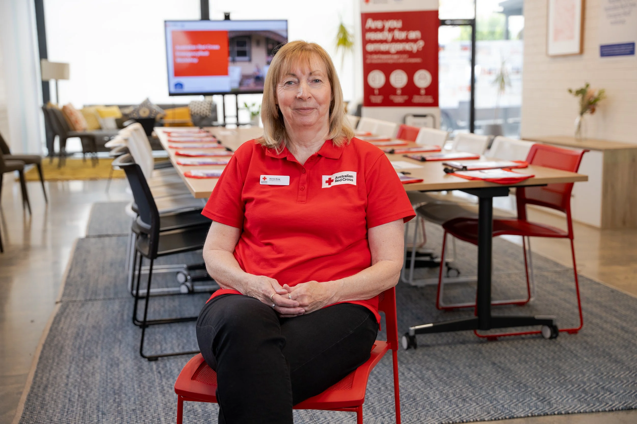 A woman sitting on a red chair in a conference room, wearing a red polo shirt with a Red Cross badge.