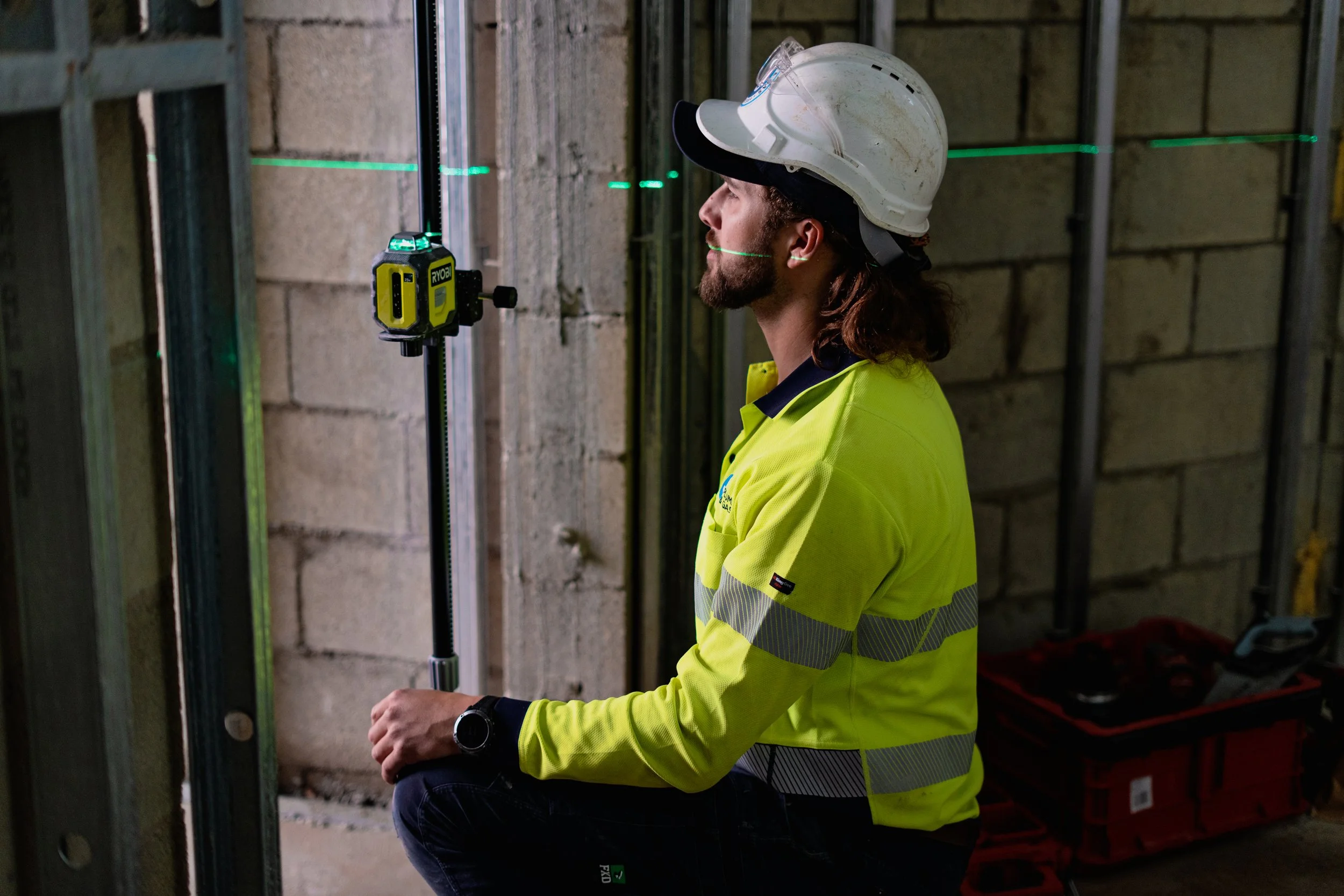 A construction worker in a yellow safety vest and white hard hat kneeling inside a building under construction, using a laser level to measure.