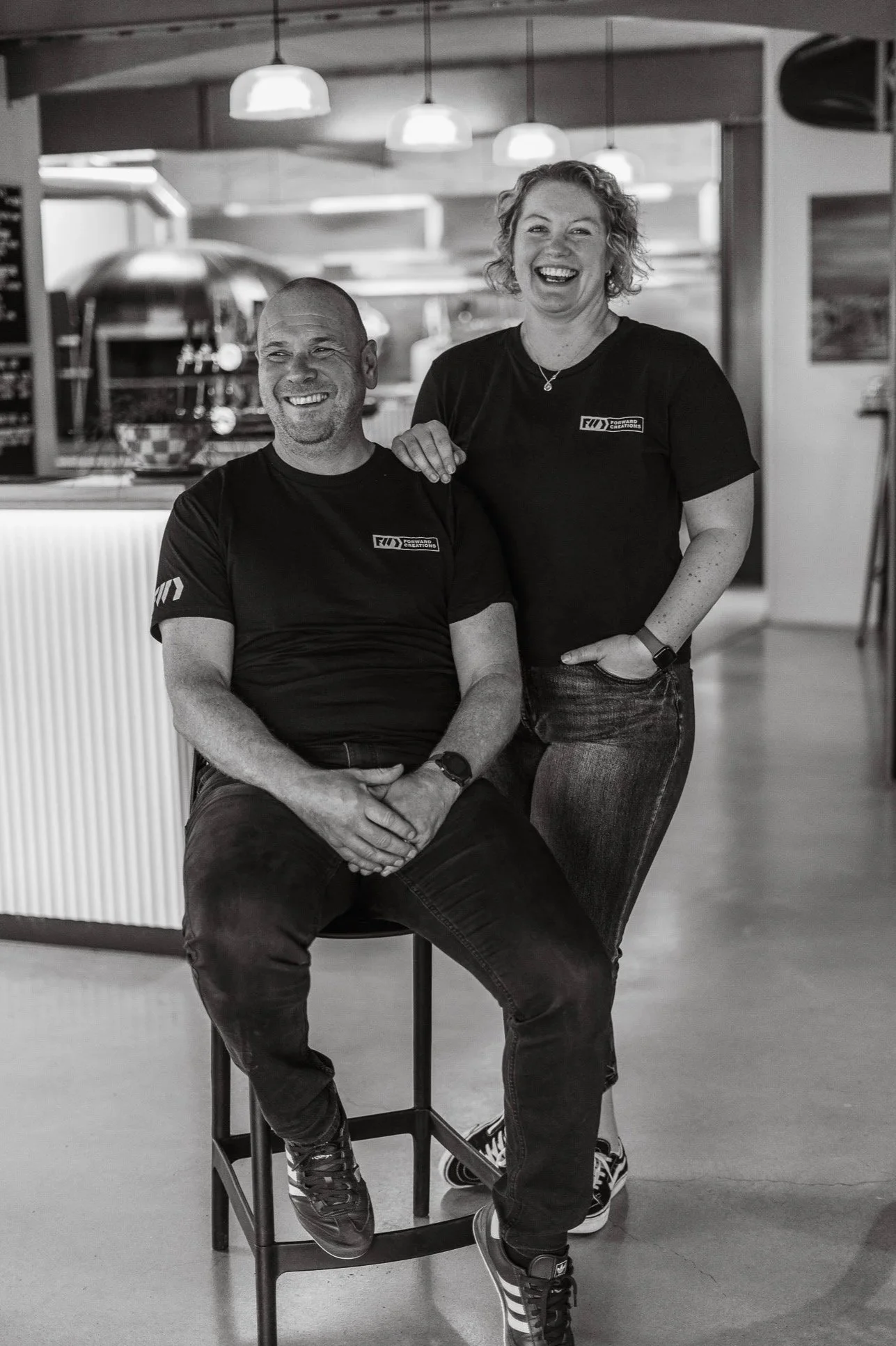 A black-and-white photo of a man sitting on a stool and a woman standing beside him, both smiling in a cafe or restaurant setting.