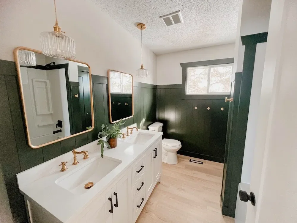 Bathroom with dual sinks, mirrors, gold fixtures, green wainscoting, and wood flooring.