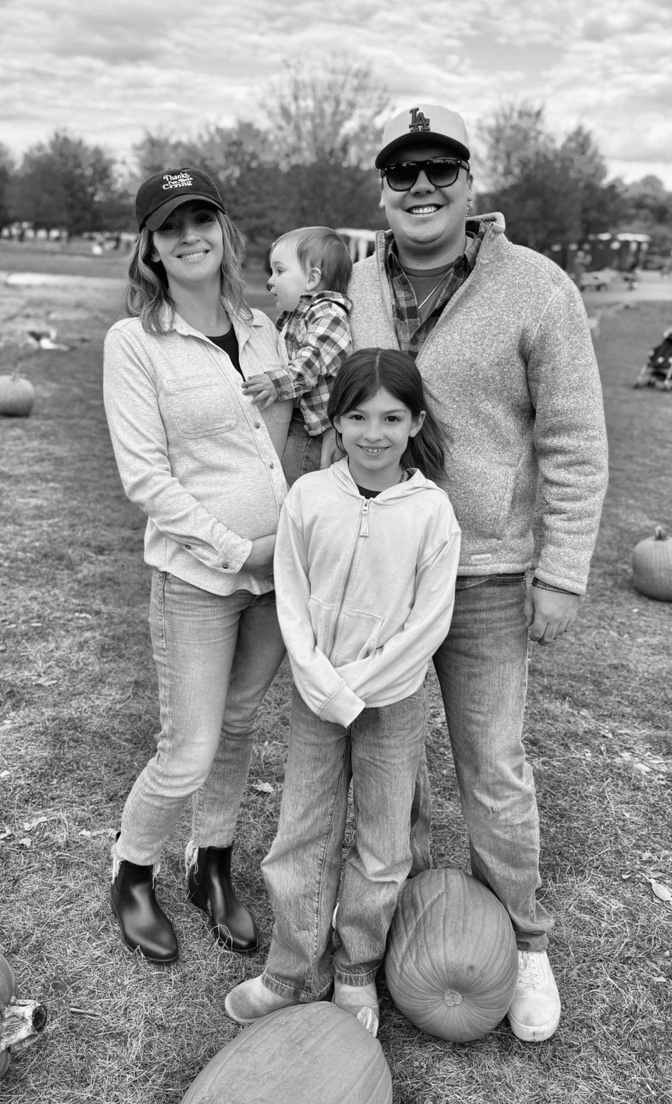 A family of four standing outdoors at a pumpkin patch, with pumpkins on the ground, in black and white.