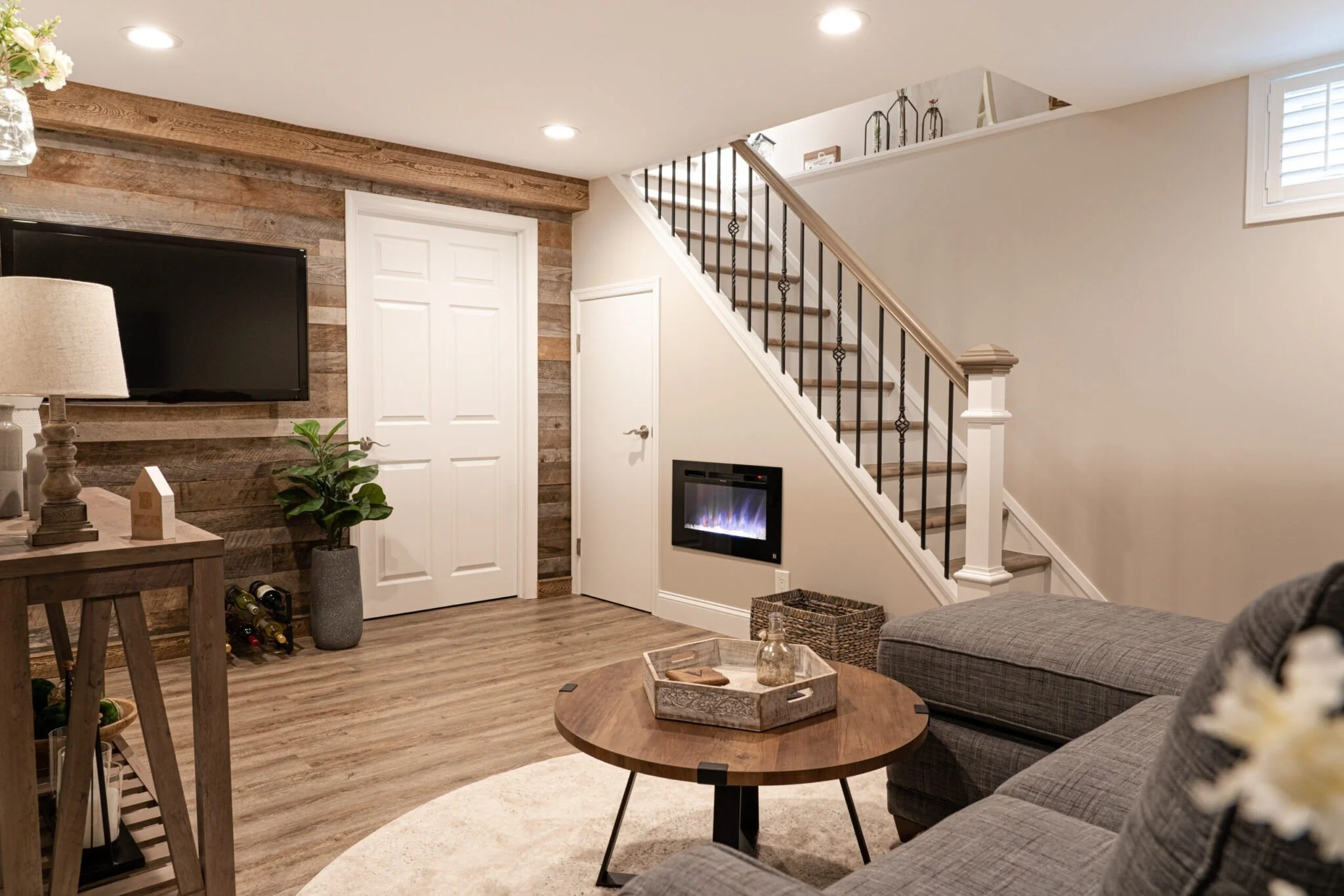 Living room with a gray sofa, wooden coffee table, fireplace, TV mounted on a wood-paneled wall, staircase with black iron balusters, a potted plant, and beige walls.