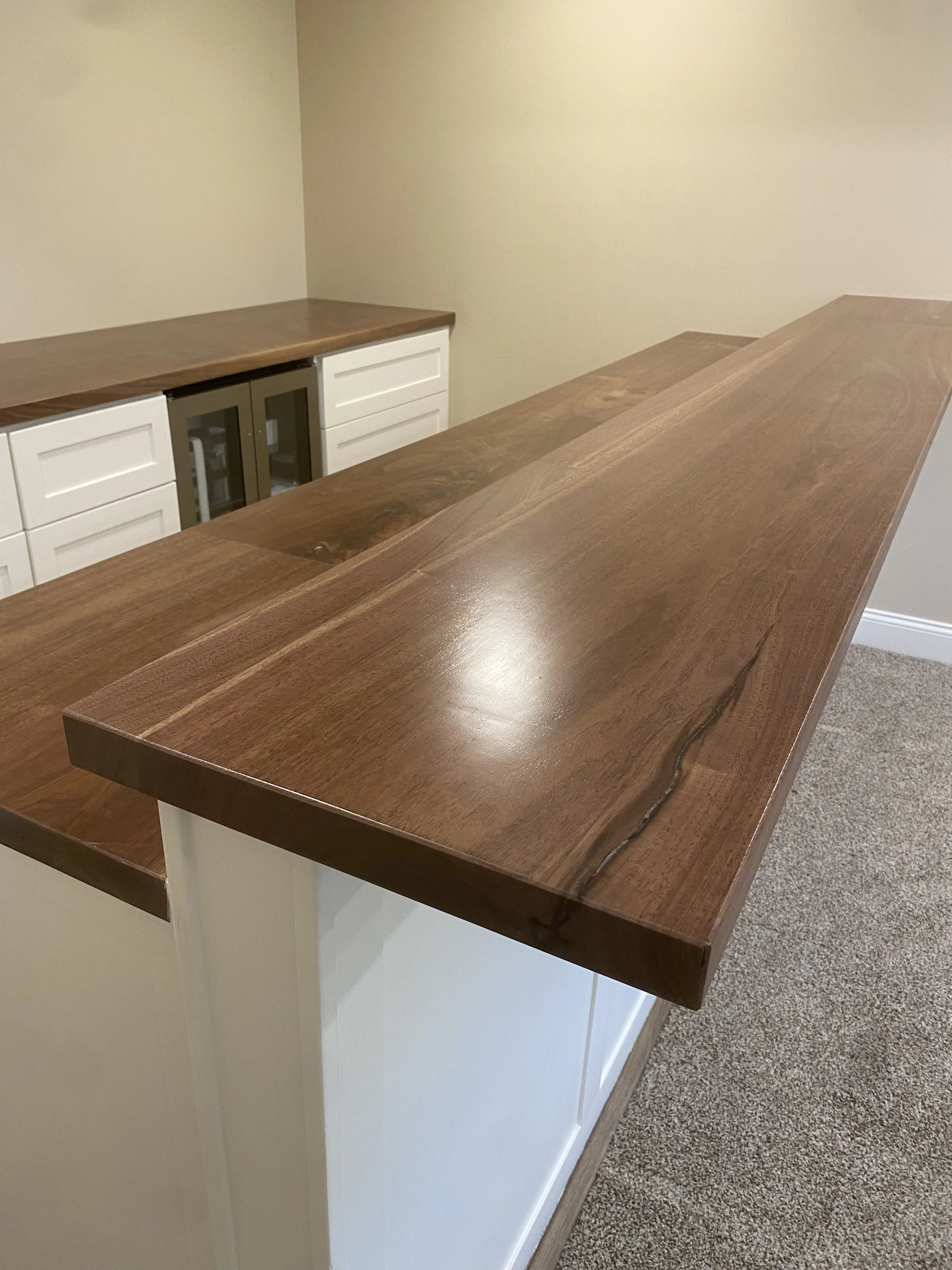 Close-up of a polished wooden countertop or bar top with natural wood grain and a live edge, with a beige wall and part of a white cabinet in the background.