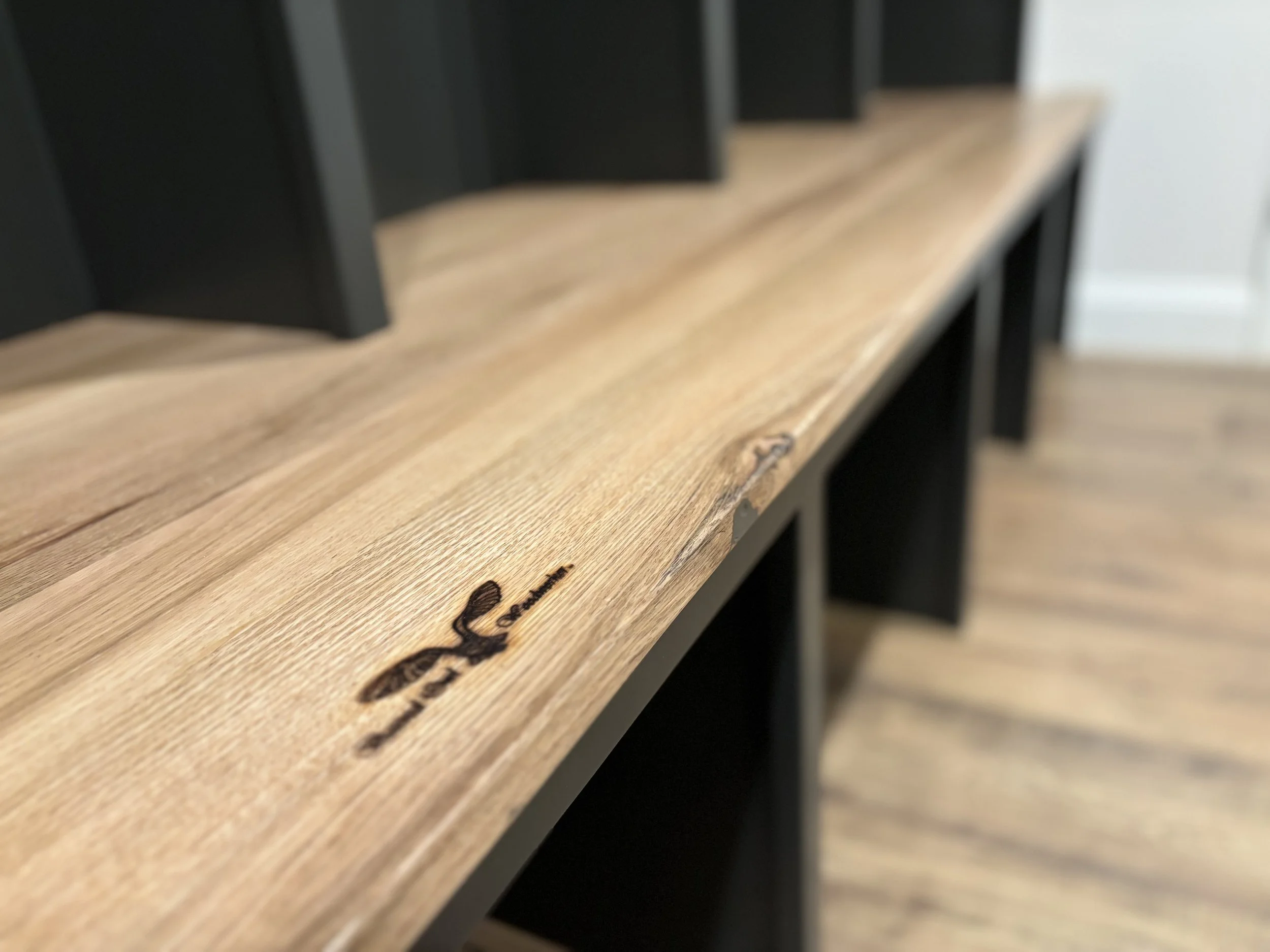 Close-up of a wooden shelf with a burned-in logo and text reading 'Defiant' in black, against a black shelf and wooden floor background.