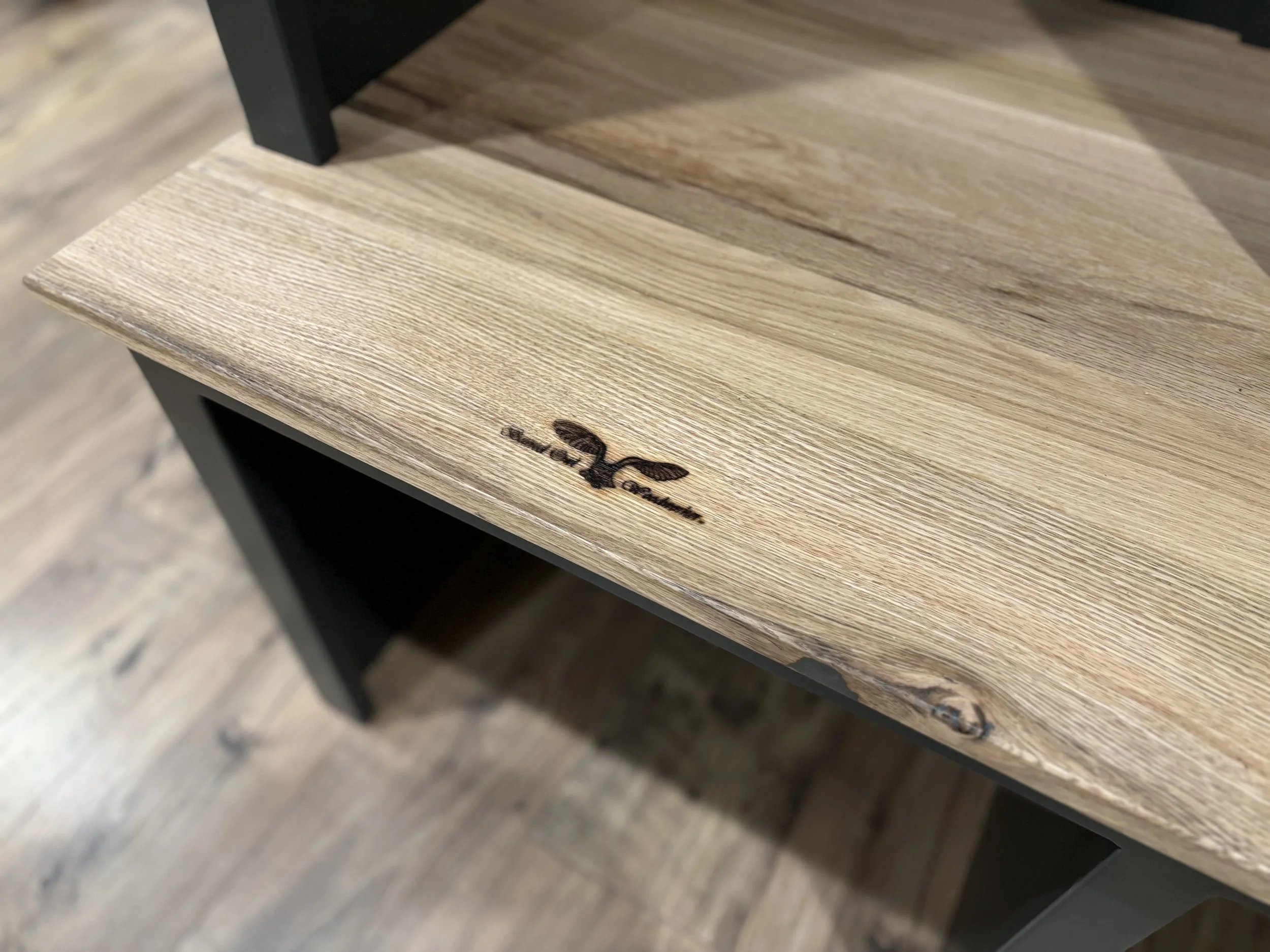 Close-up of a wooden tabletop with a burned logo or signature and black furniture legs.