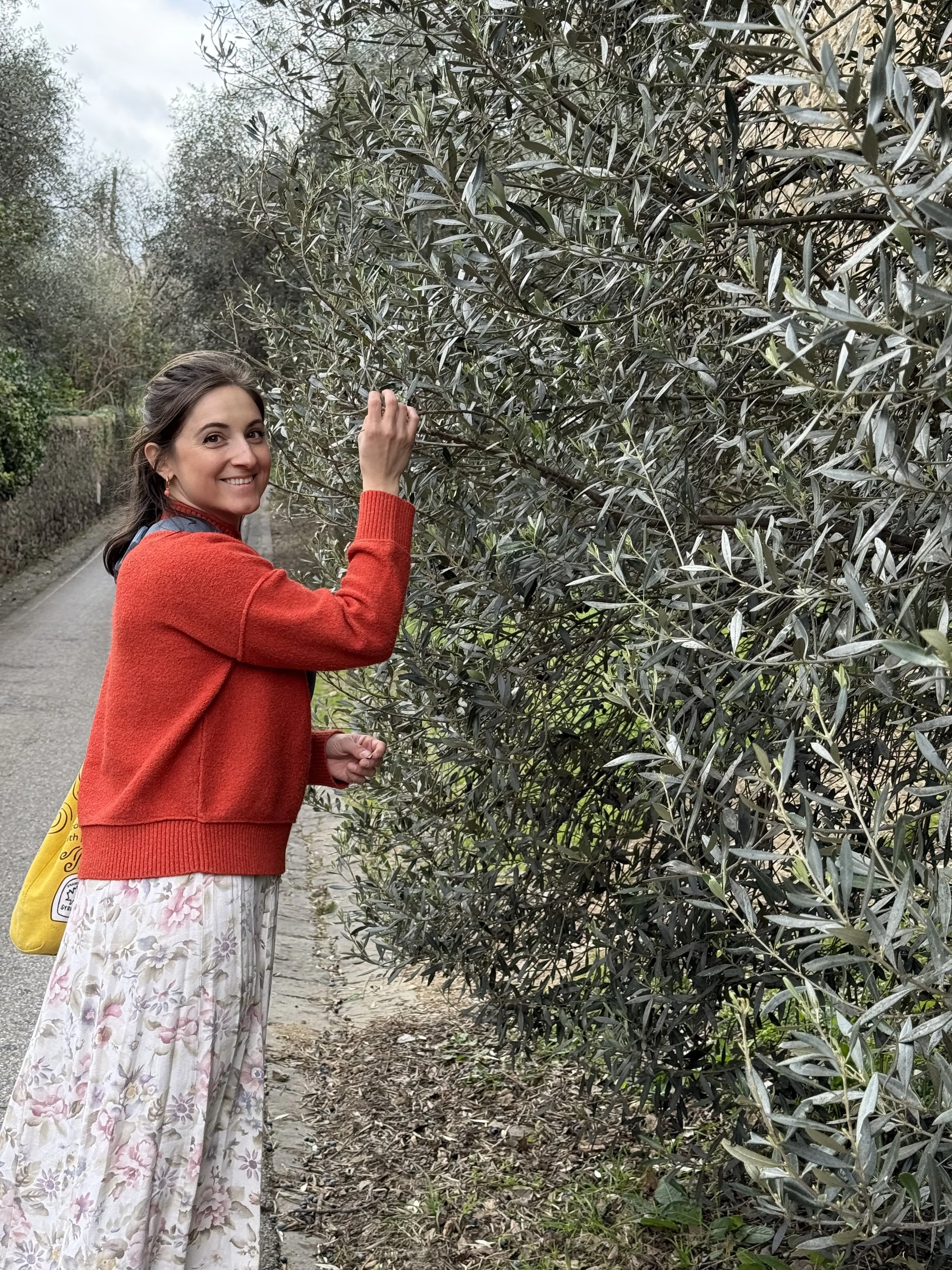 A woman in a red sweater and a floral skirt is standing by a hedge and picking a leaf from the plant.