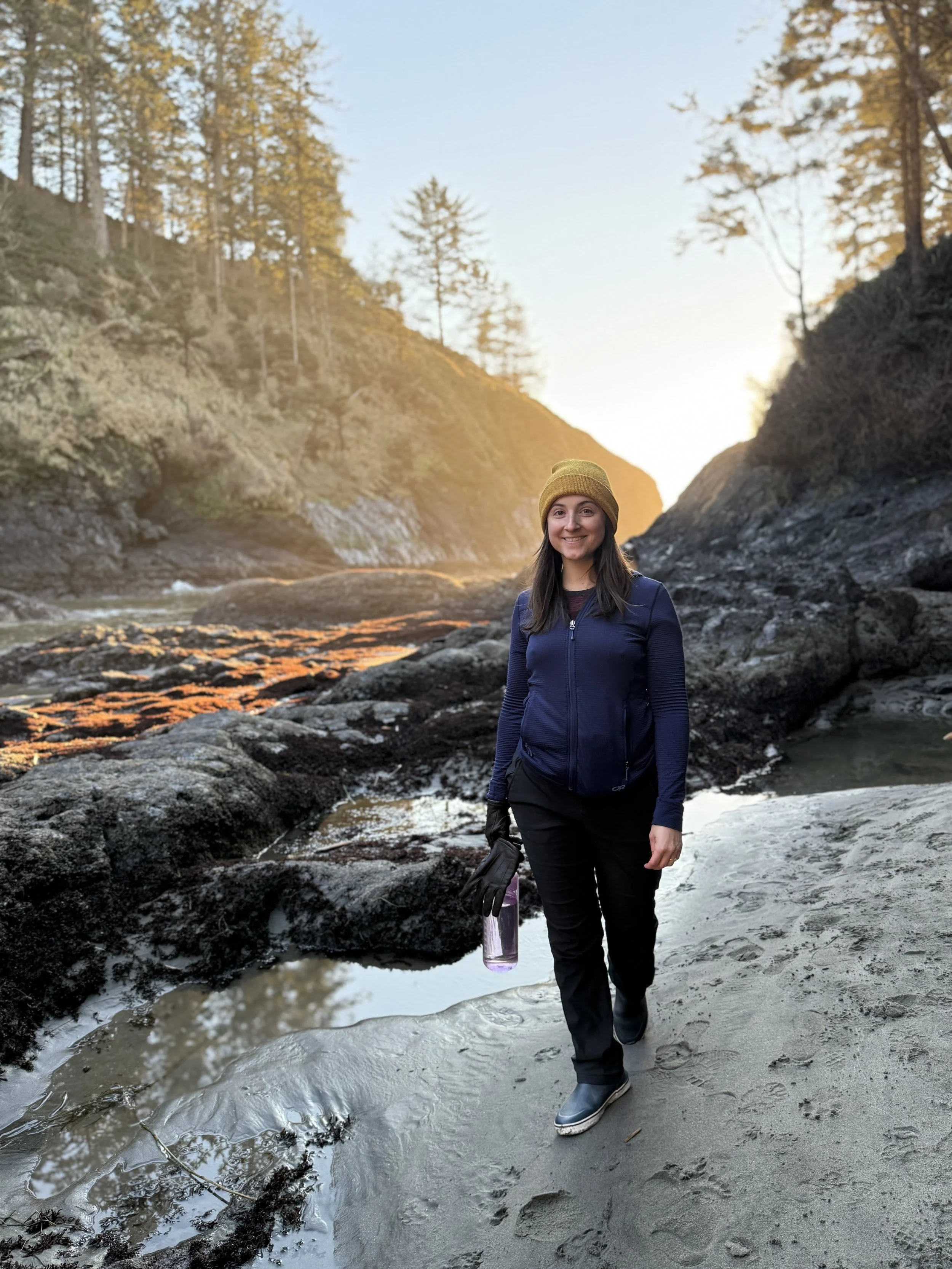 A woman walking on a rocky beach at sunset, surrounded by trees on cliffs.