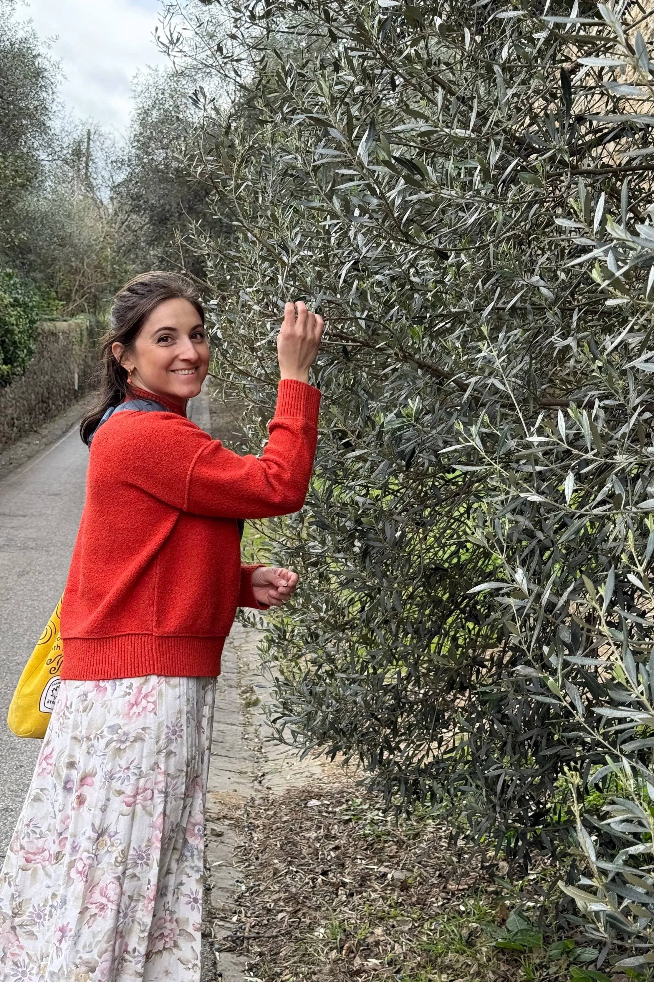 Monique, Explore Amore Founder, admiring an olive bush in Italy, wearing a floral dress and red sweater.