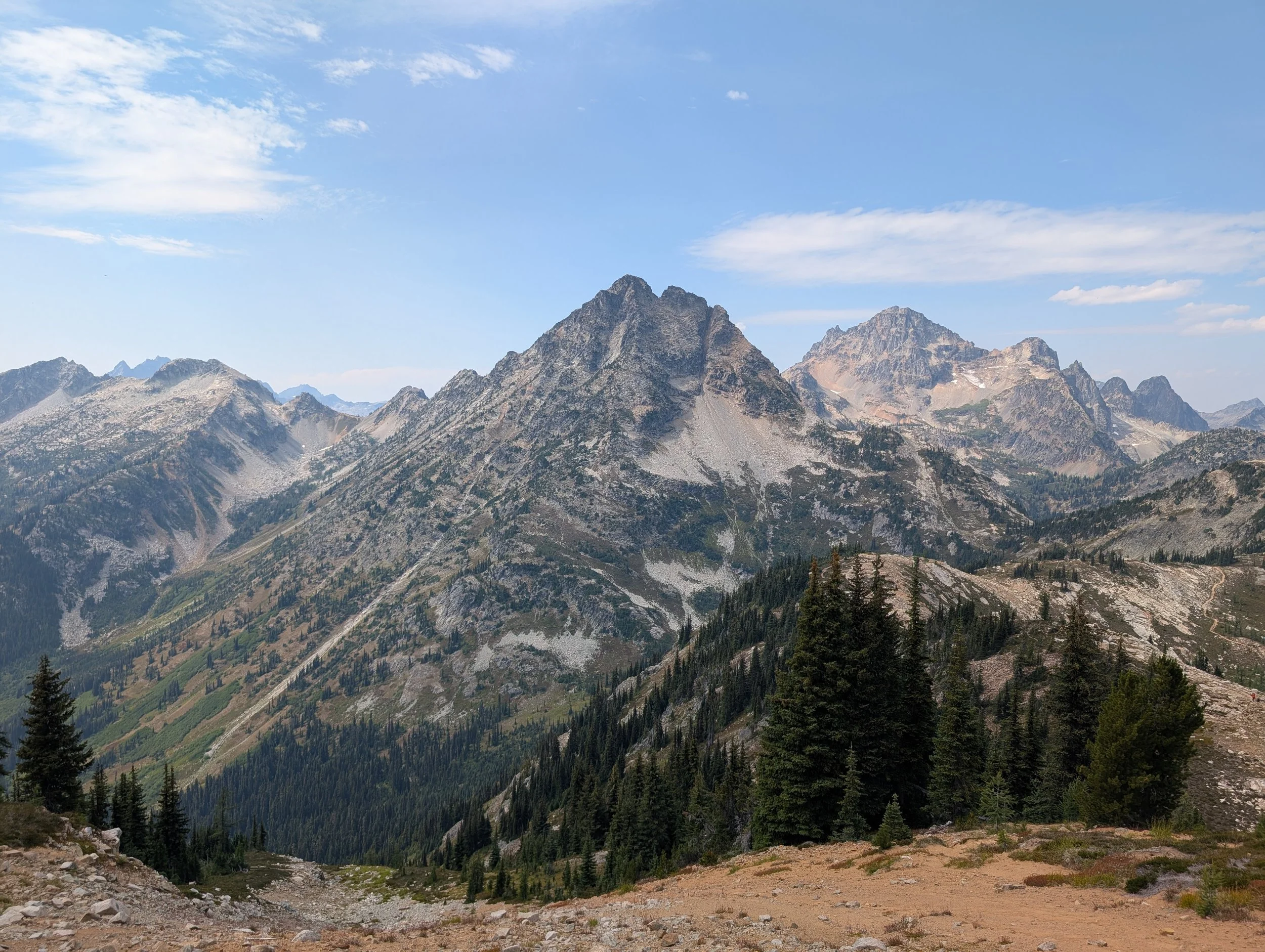 Mountain landscape with rugged peaks, green forested slopes, and a blue sky with scattered clouds.