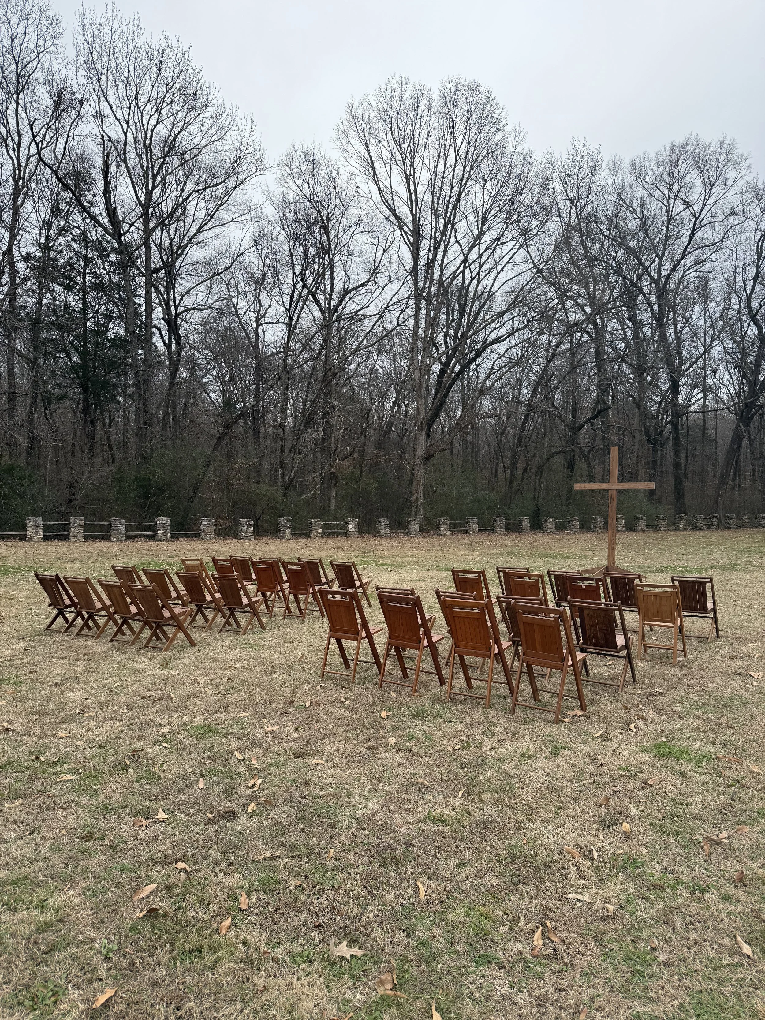 Outdoor scene with wooden chairs arranged in a semi-circle around a large wooden cross, set on a grassy field with trees in the background on a cloudy day.