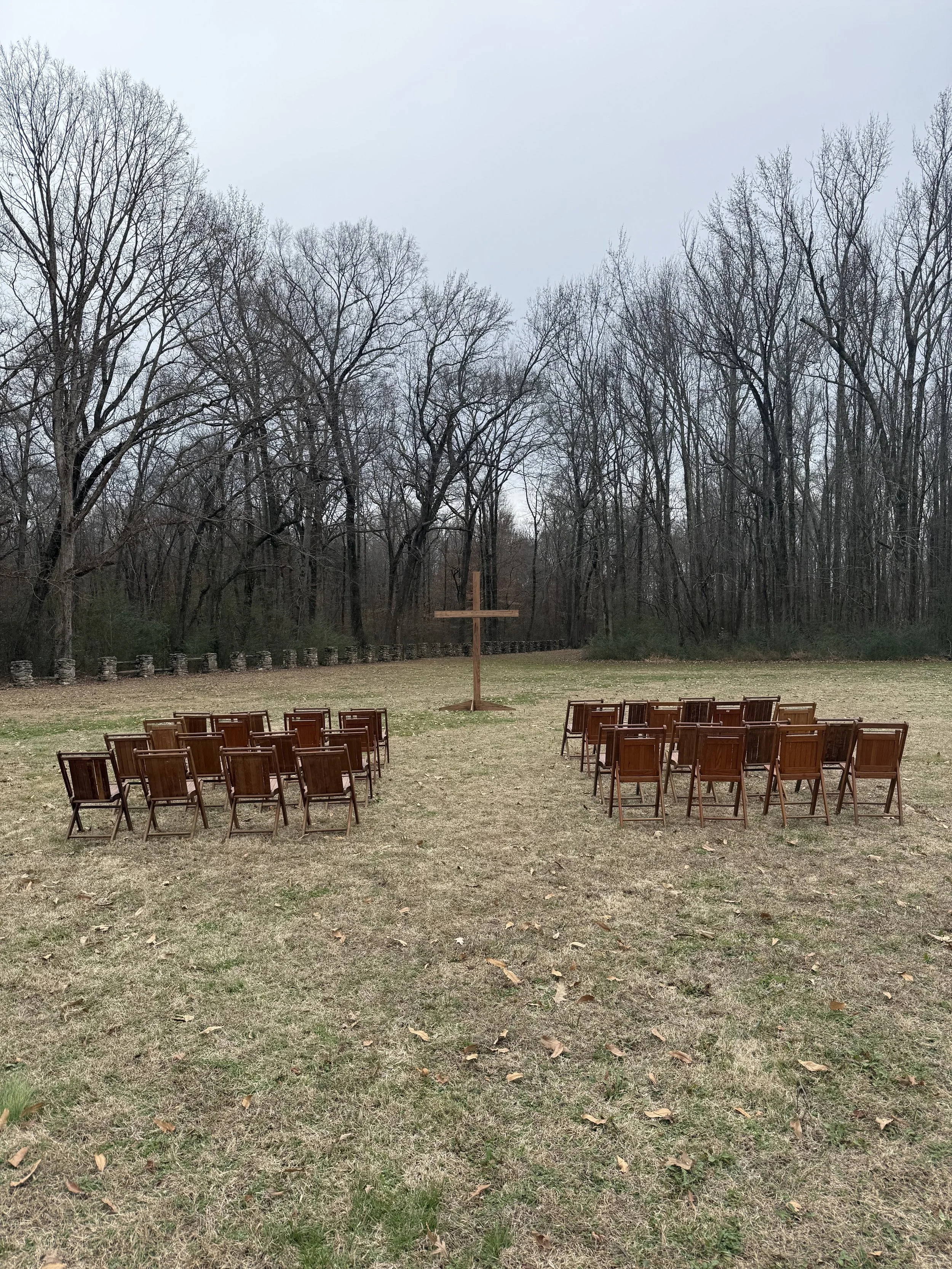 Chairs arranged in two groups facing a wooden cross in an outdoor setting with trees in the background.