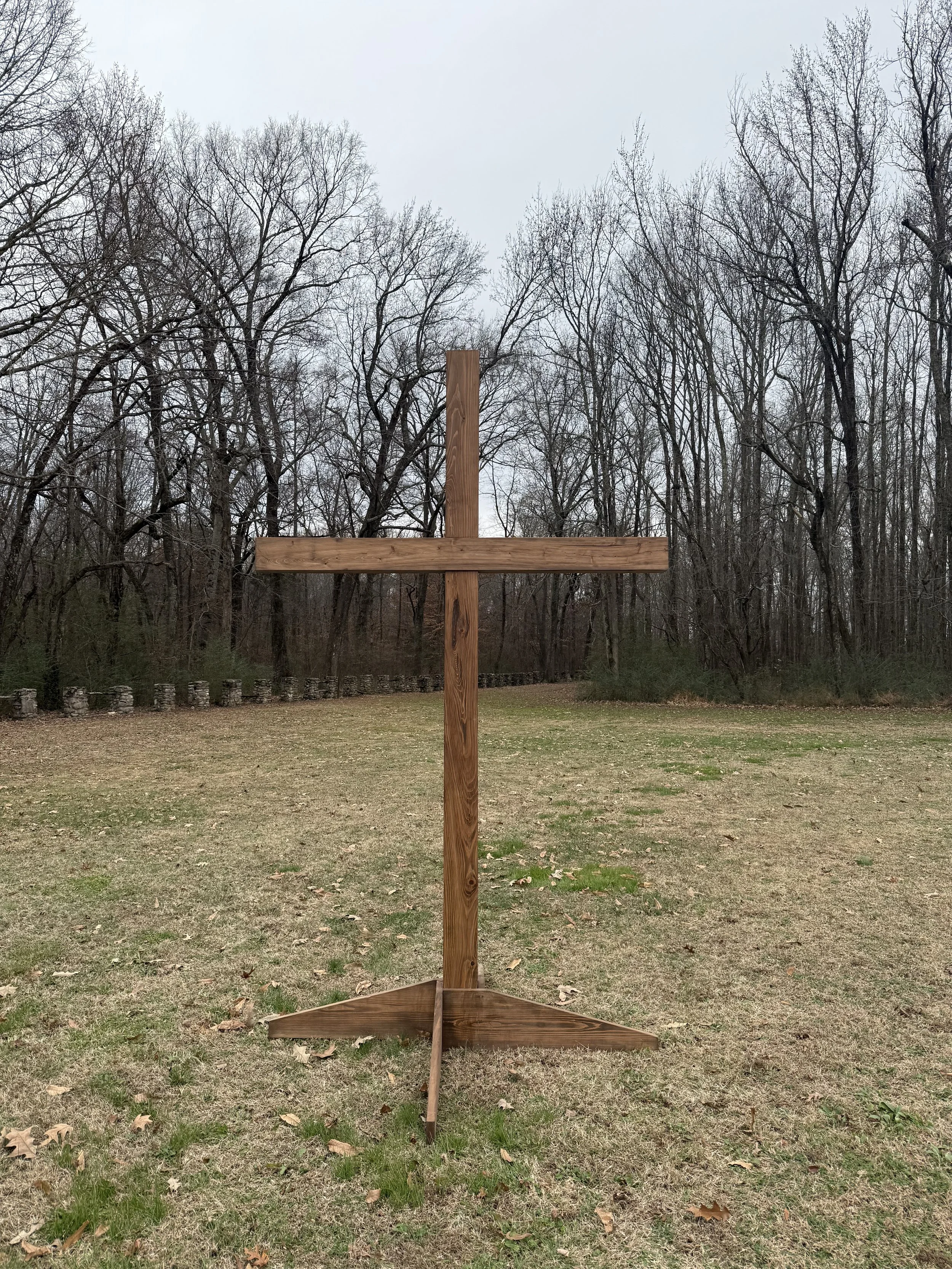 A wooden cross standing outdoors on a grassy field with trees in the background and a cloudy sky overhead.
