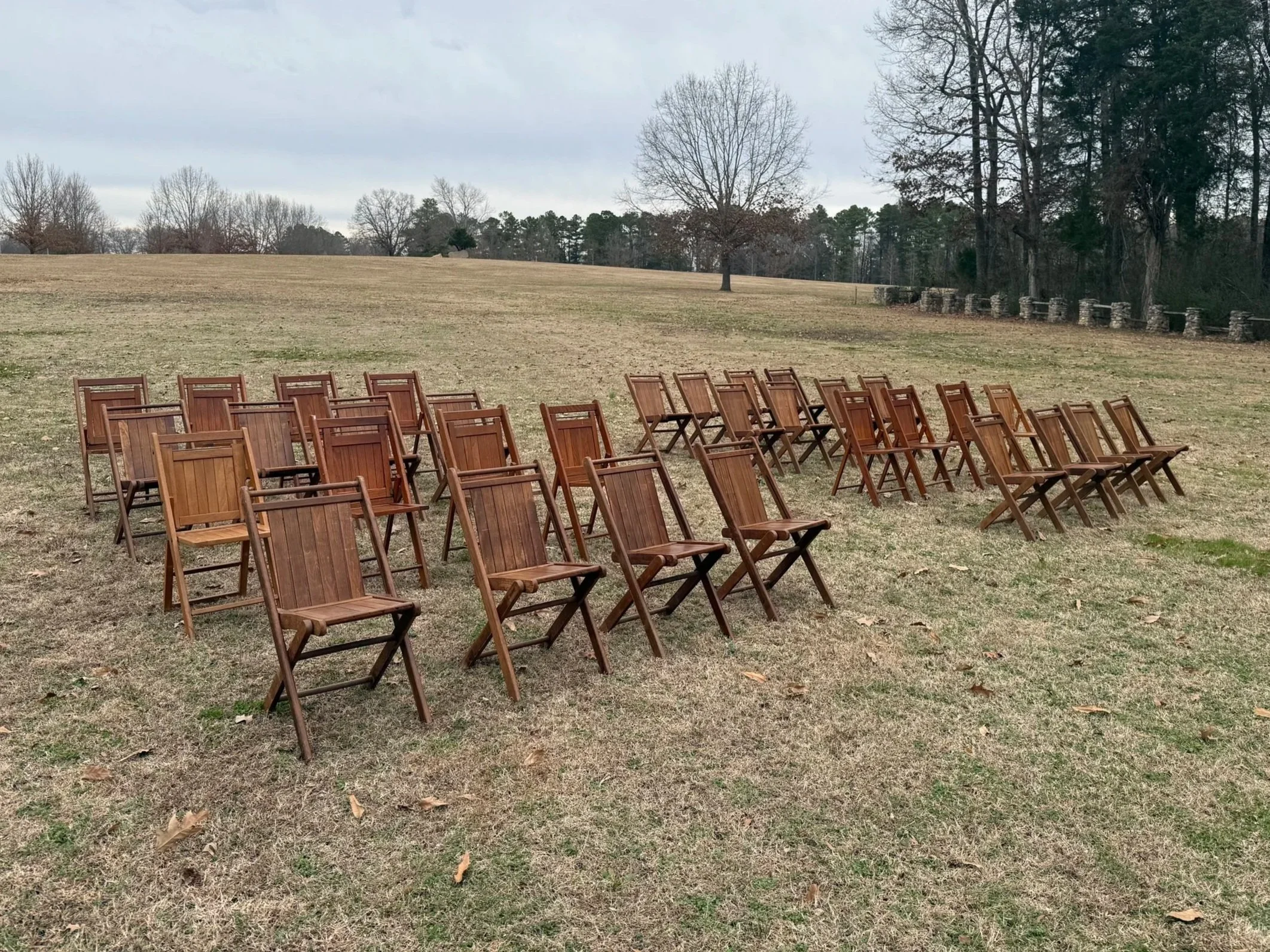 Arranged wooden chairs on a grassy field with trees and cloudy sky in the background.
