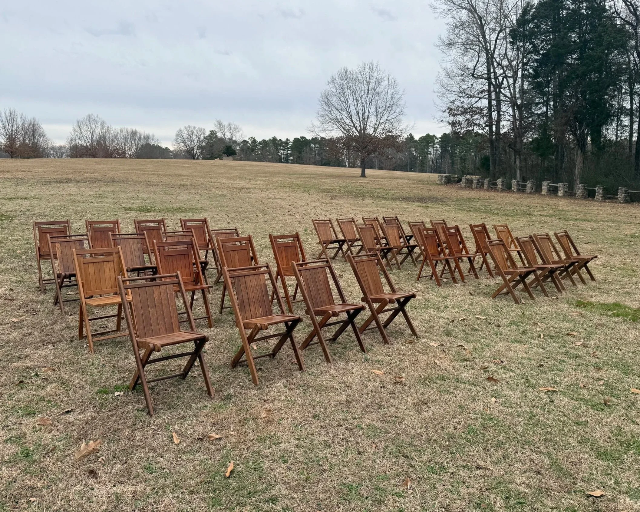 Rows of empty wooden folding chairs arranged outdoors on grass, set against a backdrop of trees and cloudy sky, likely for an event or gathering.