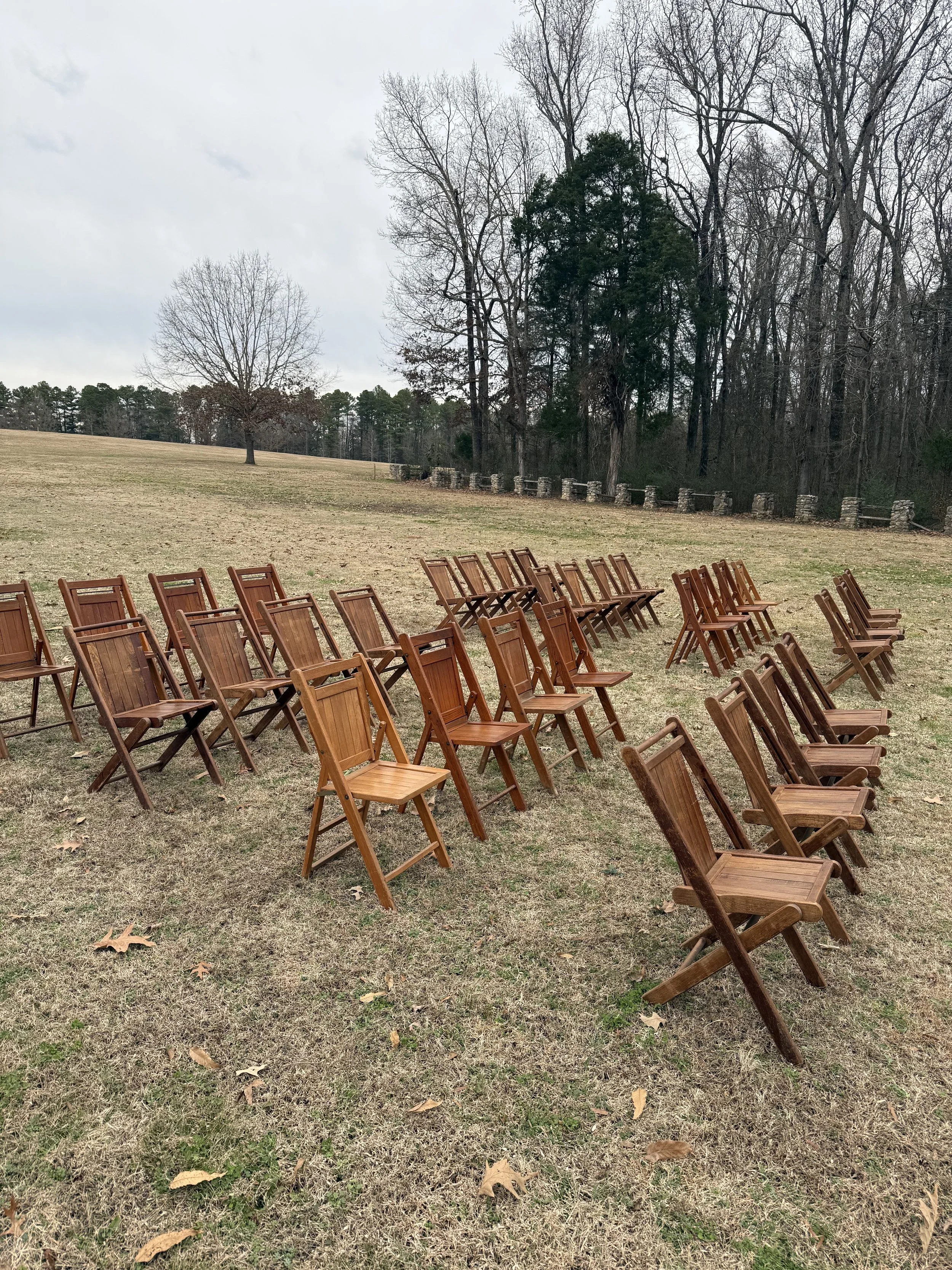 Multiple wooden chairs arranged in rows facing away towards a grassy field with trees and a stone wall in the background, under an overcast sky.
