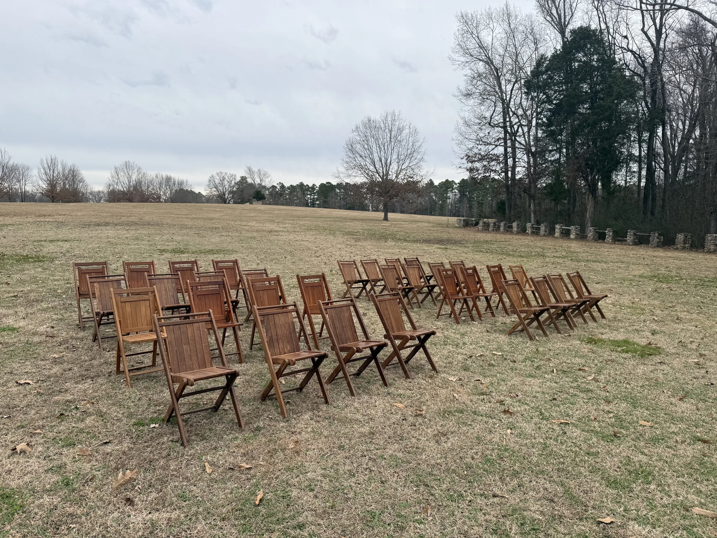Multiple wooden chairs arranged outdoors on a grassy field on a cloudy day, with trees in the background.