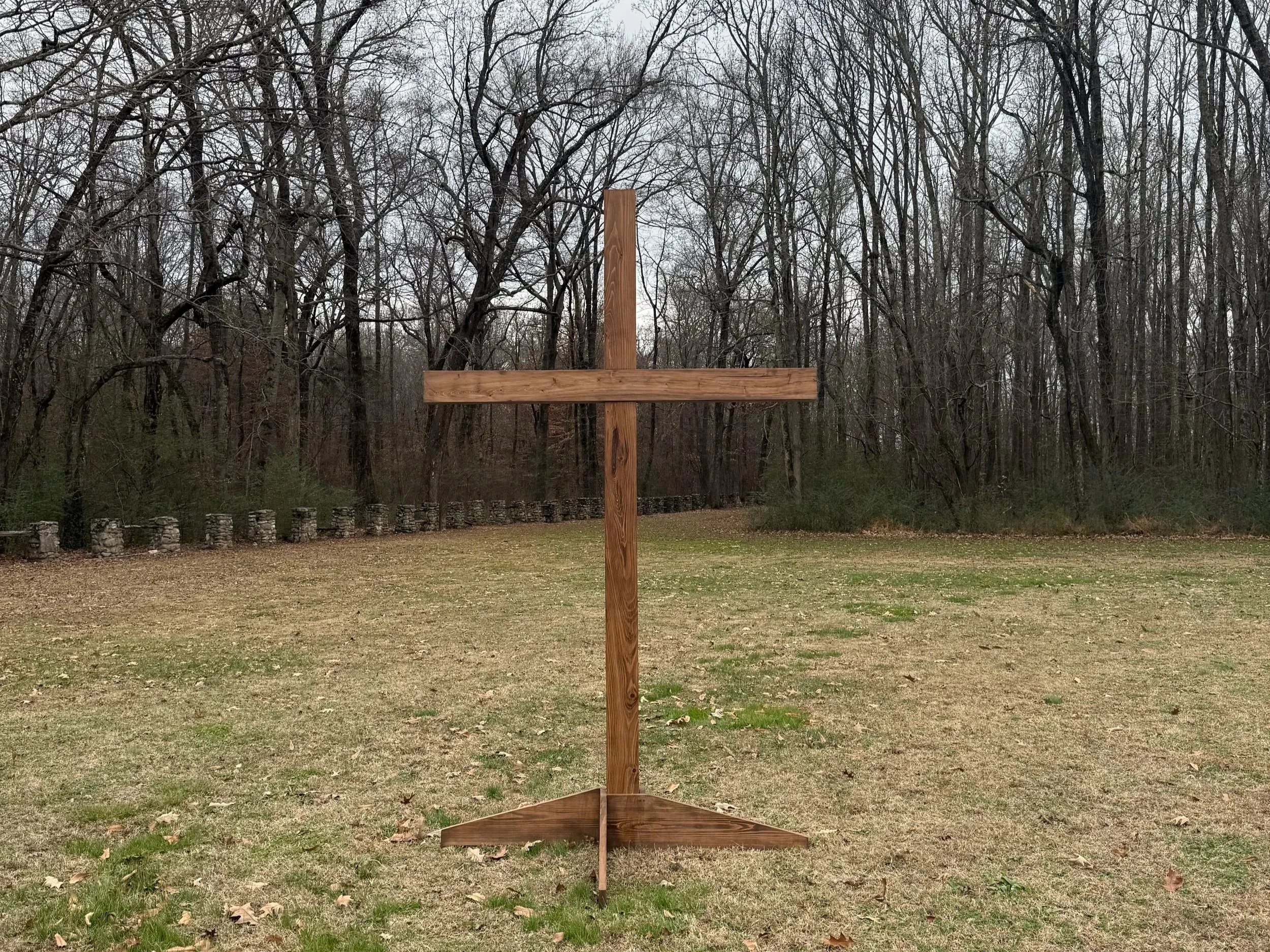 A large wooden cross standing on a grassy field with a forest of leafless trees in the background.