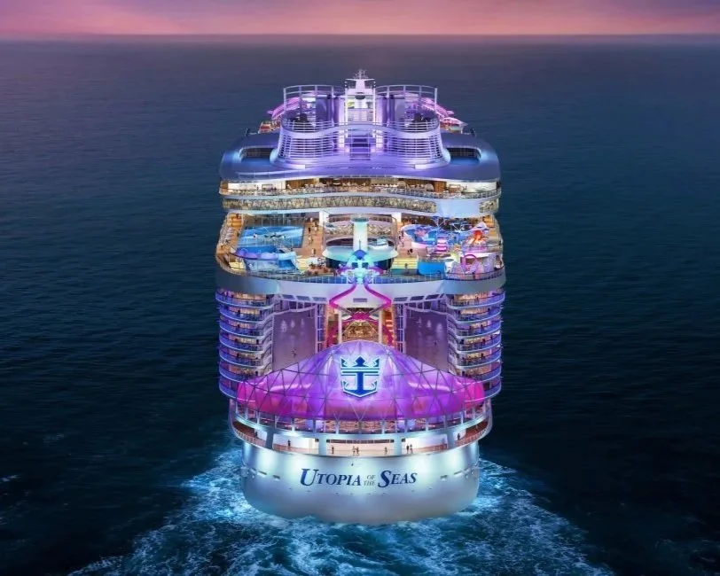 A large cruise ship named Utopia of the Seas sailing on the ocean at sunset, with illuminated decks and various recreational areas visible.
