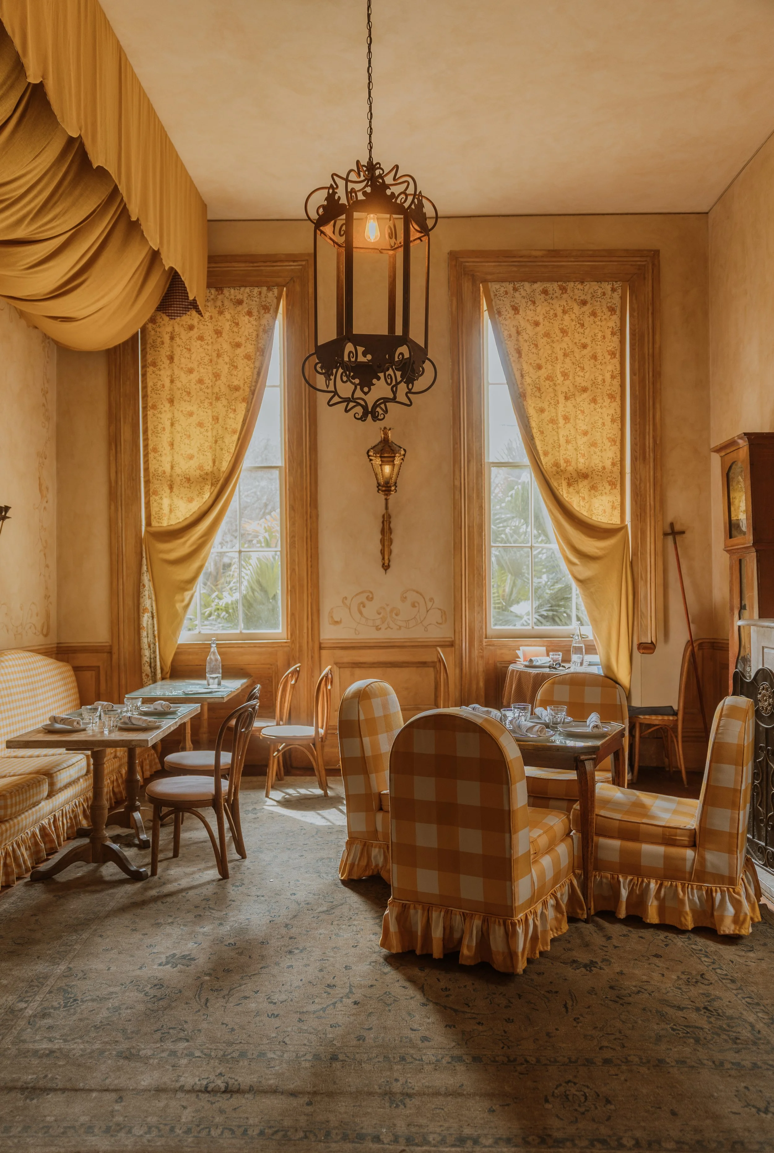 Elegant dining room with yellow checkered curtains and chairs, wooden tables set with glassware, and a vintage chandelier hanging from the ceiling.