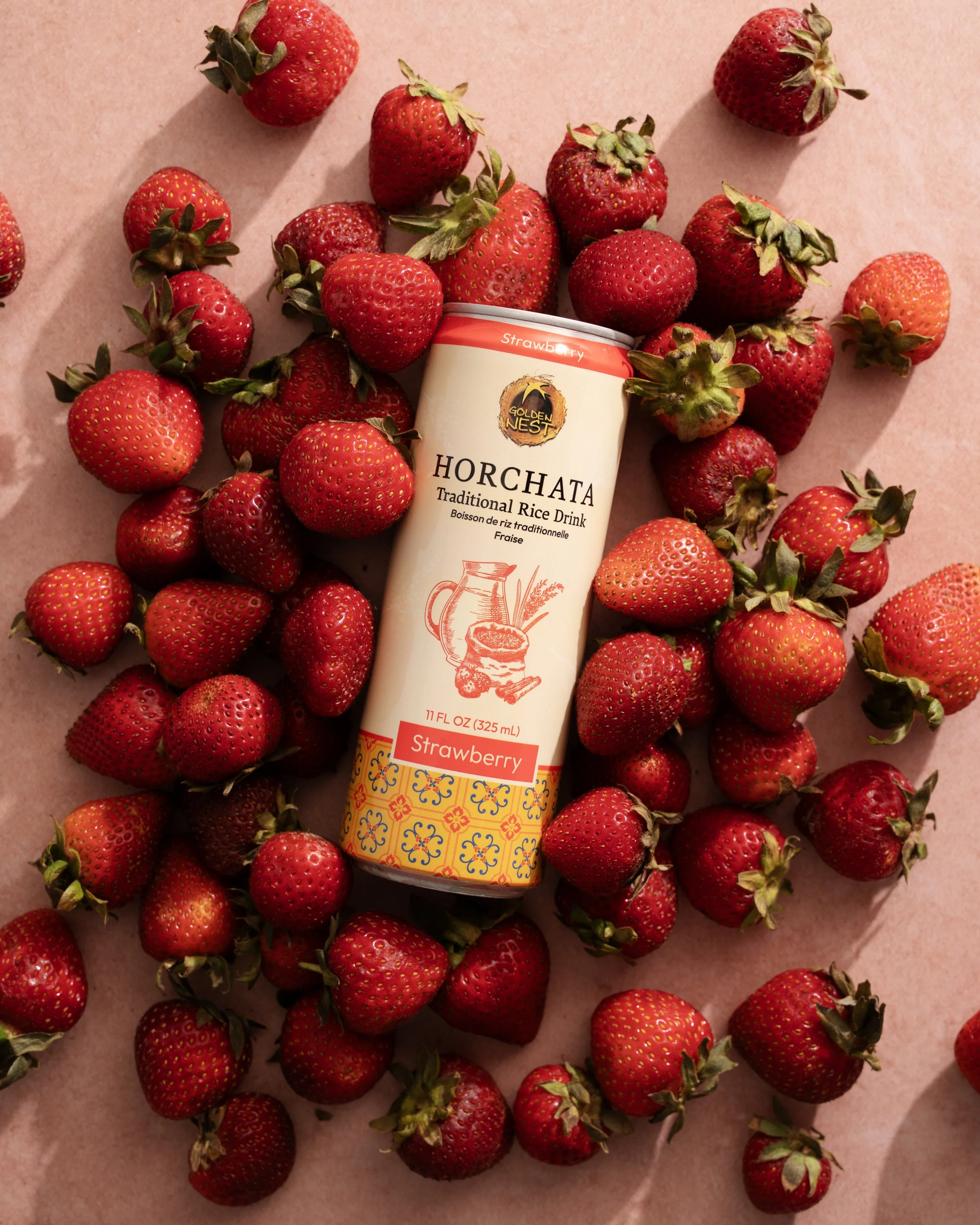 A can of Golden Nest Horchata surrounded by fresh strawberries on a pink surface.