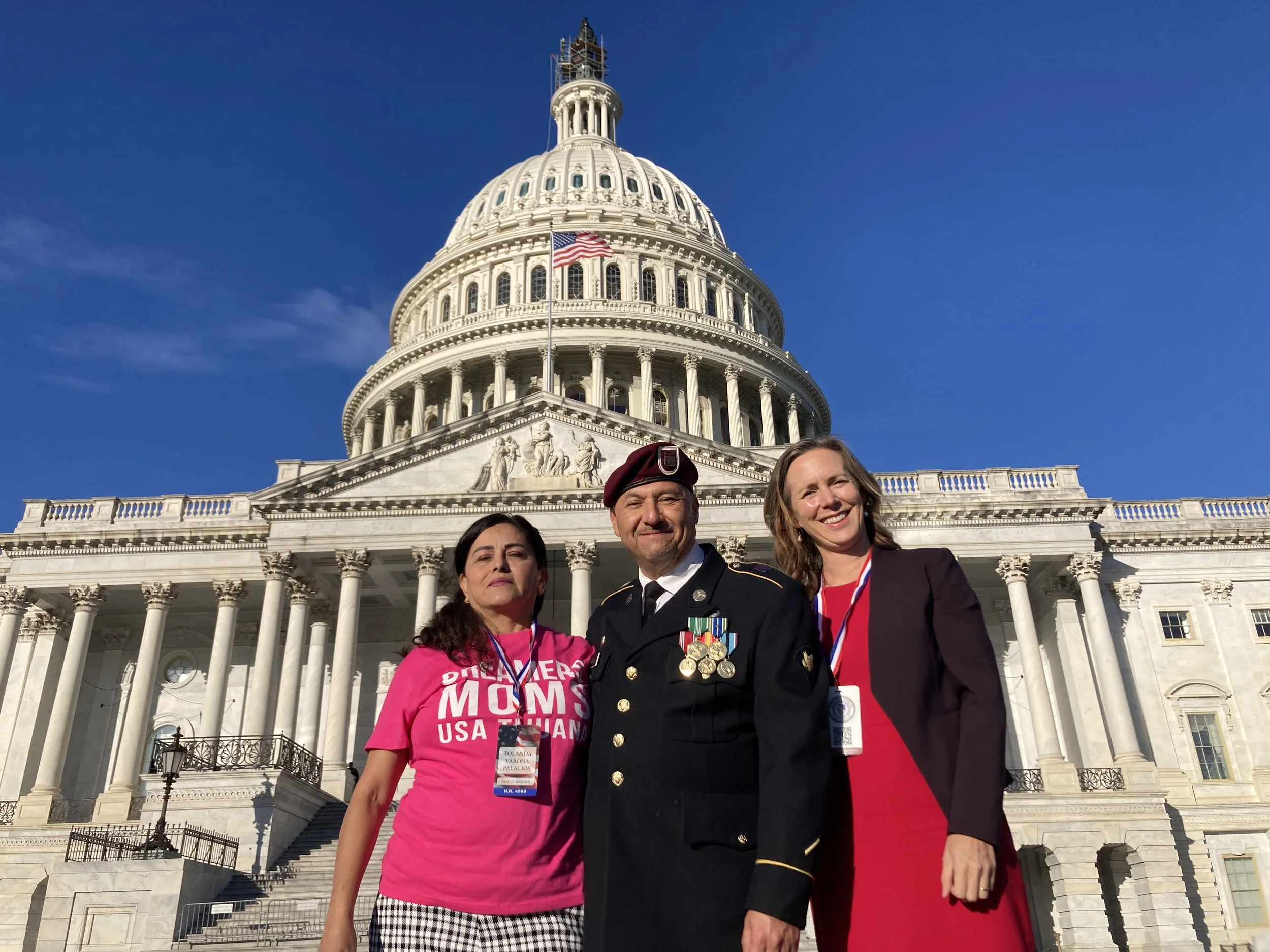 Three people standing in front of the U.S. Capitol building. One woman wearing a pink shirt that says 'FIGHTING FOR OUR MOMS' and checkered pants, a man in a military uniform with medals, and another woman in a red dress with a dark blazer. All three are smiling and wearing lanyards with badges, with the Capitol dome in the background against a clear blue sky.
