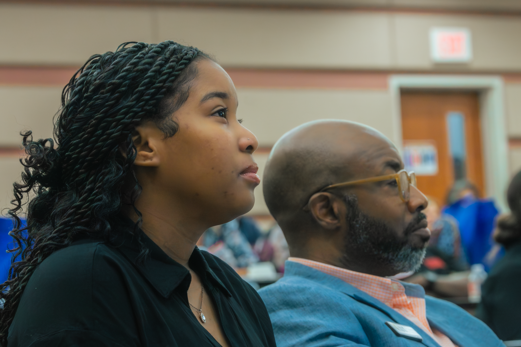 Side view of a young woman with braided dark hair and a man with glasses and a beard sitting in a conference room, listening attentively.