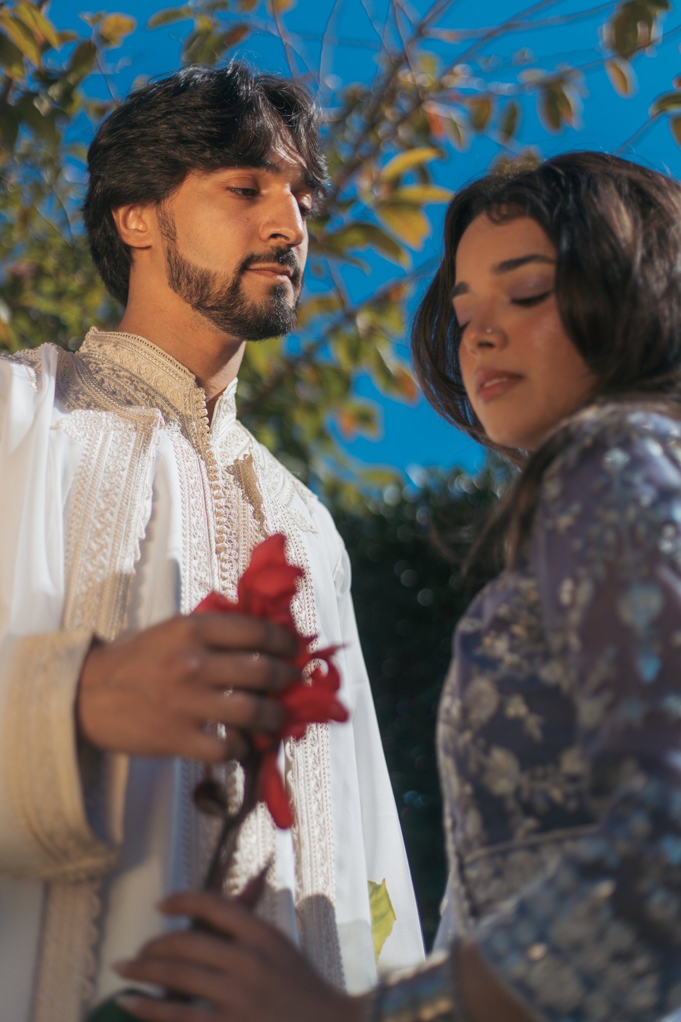 A man in traditional Indian attire holds a red flower in his hand as he looks down at a woman with dark hair in floral clothing, set outdoors with trees and a clear blue sky in the background.