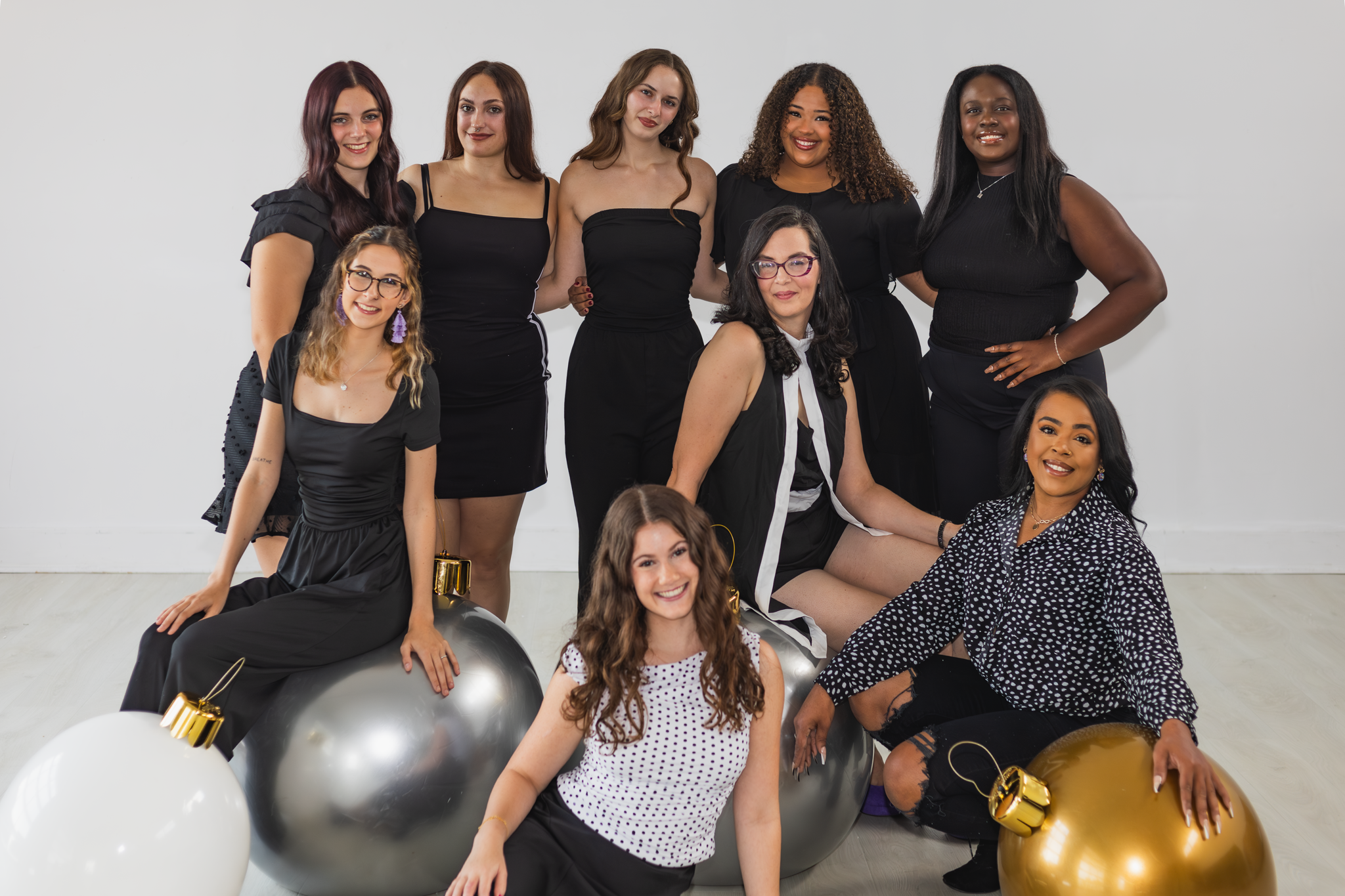 A diverse group of ten women posing together indoors with Christmas ornaments, wearing black and white outfits, smiling at the camera.