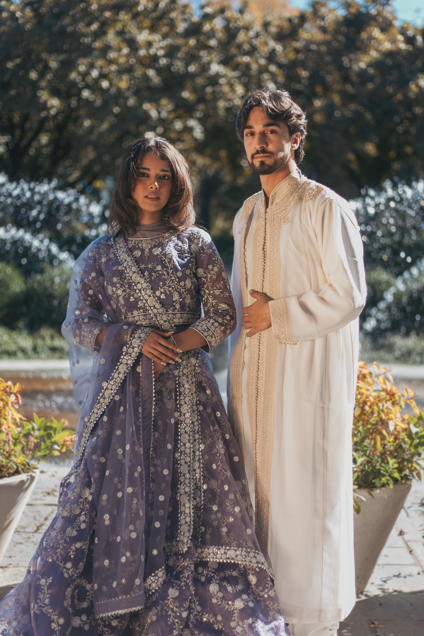 A man and woman dressed in traditional Indian attire standing outdoors with plants and trees in the background.