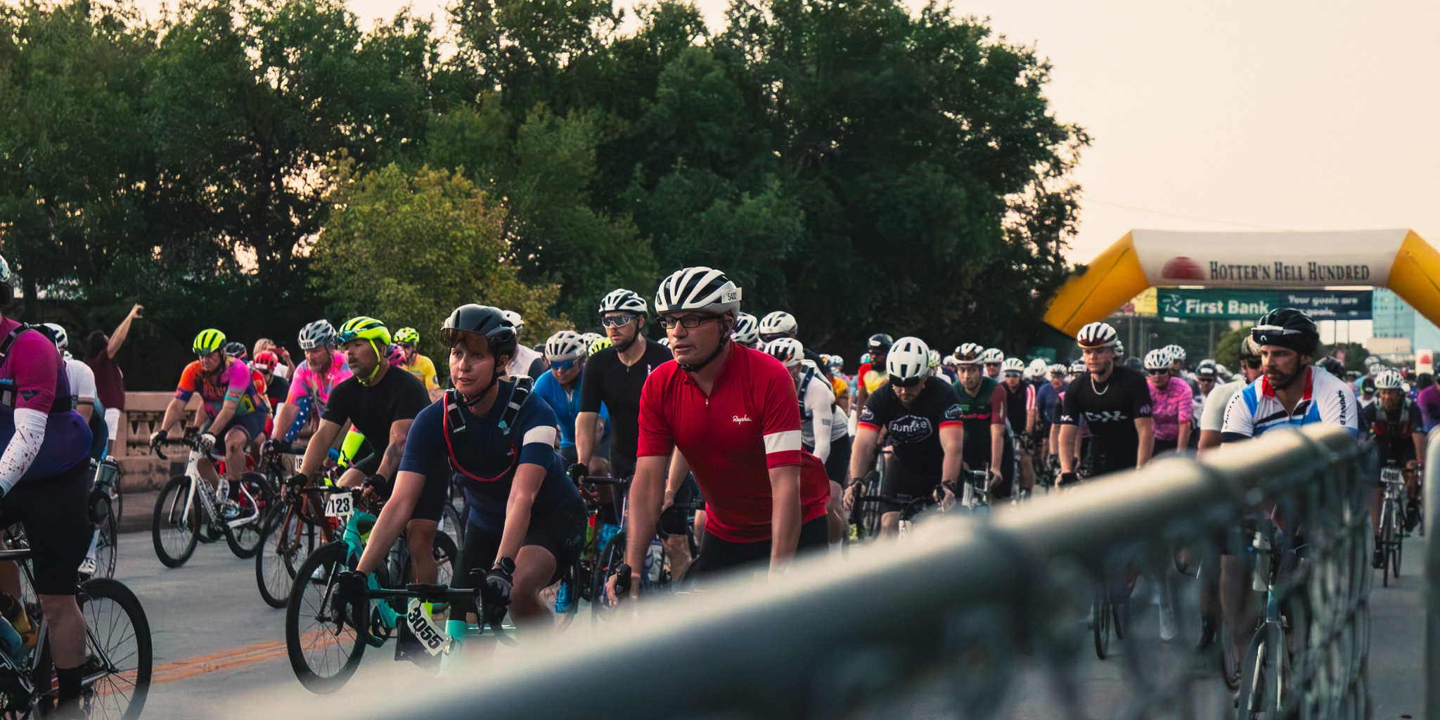 A large group of cyclists at a race start line, wearing helmets, colorful jerseys, and sunglasses, behind a safety barrier, with a yellow archway in the background over the street.