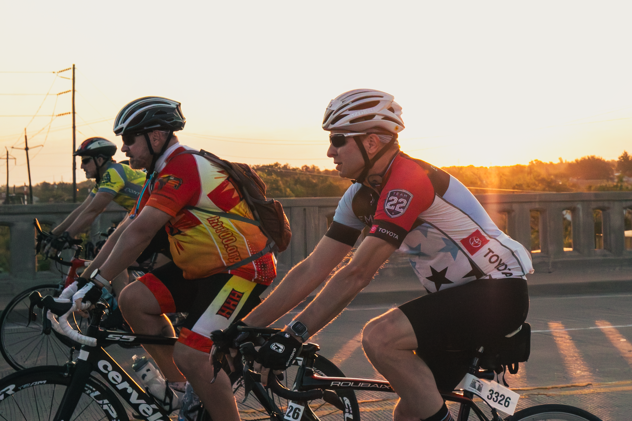 Three cyclists riding on a bridge at sunset, wearing helmets and athletic gear.