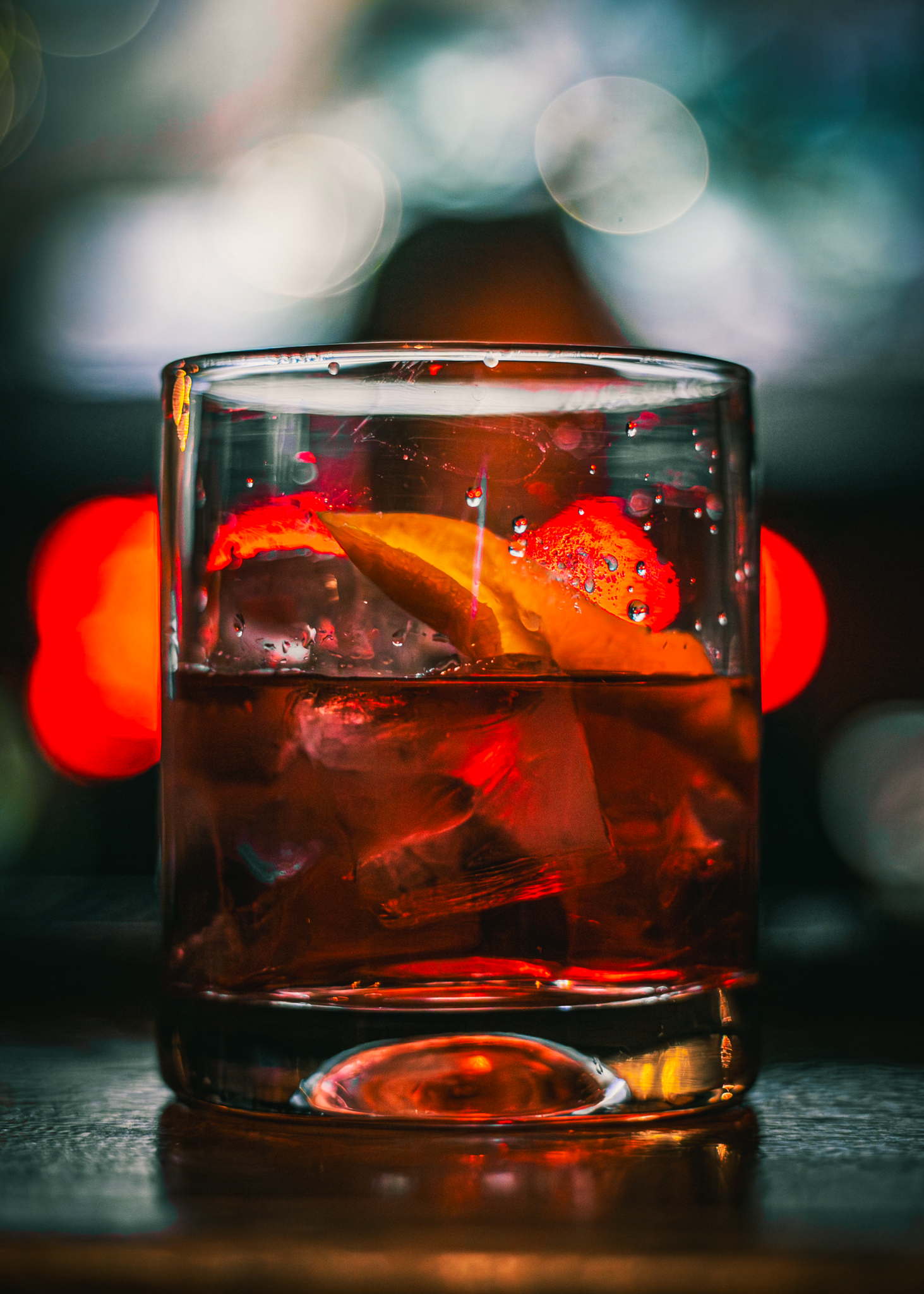 A close-up of a glass of red cocktail with ice and orange slices, set against blurred colorful background lights.