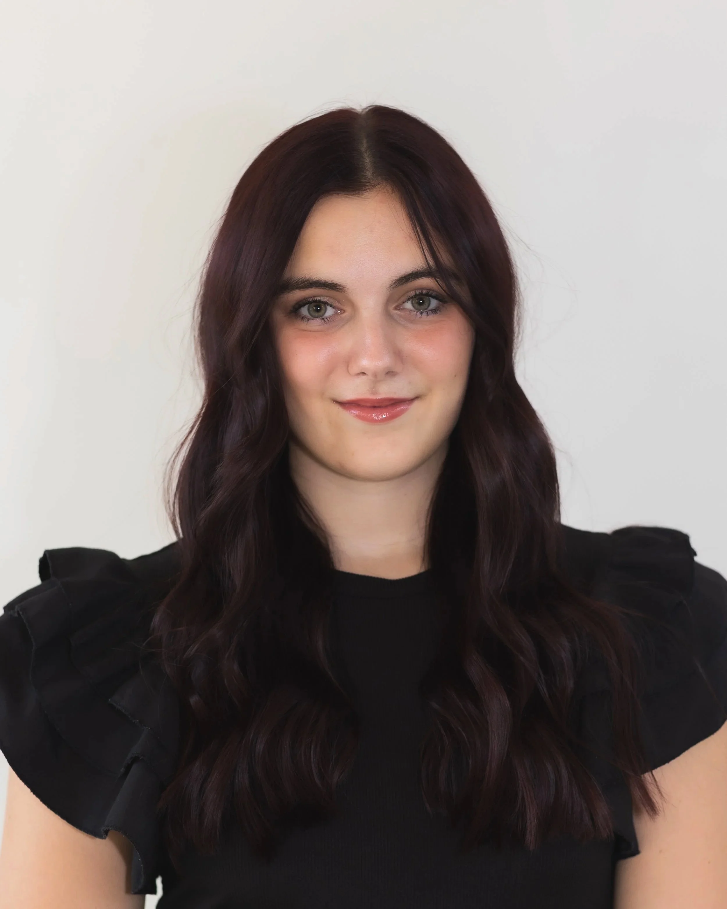 Young woman with dark wavy hair, wearing a black top with ruffled shoulders, smiling against a plain white background.