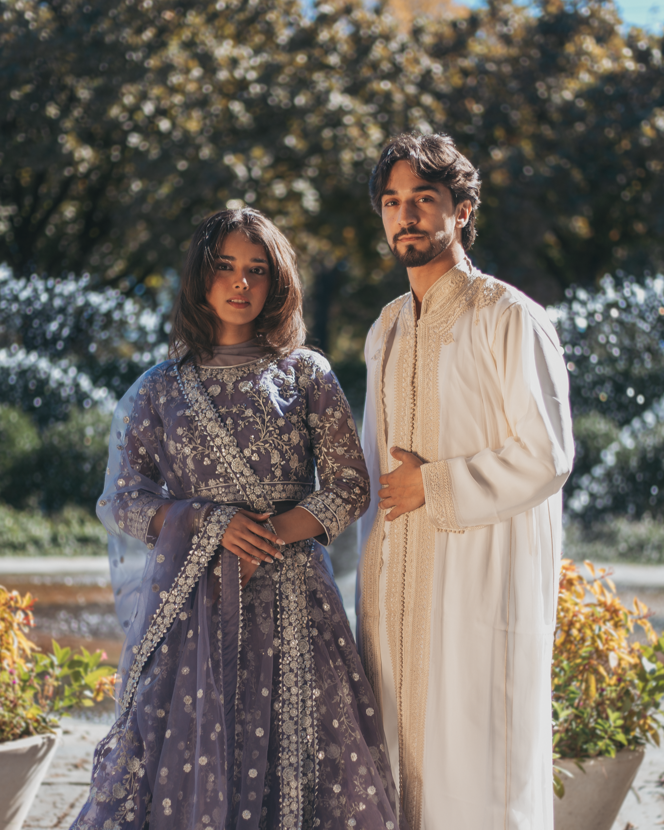 A man and woman in traditional Indian attire standing outdoors in front of a background of trees and potted plants.