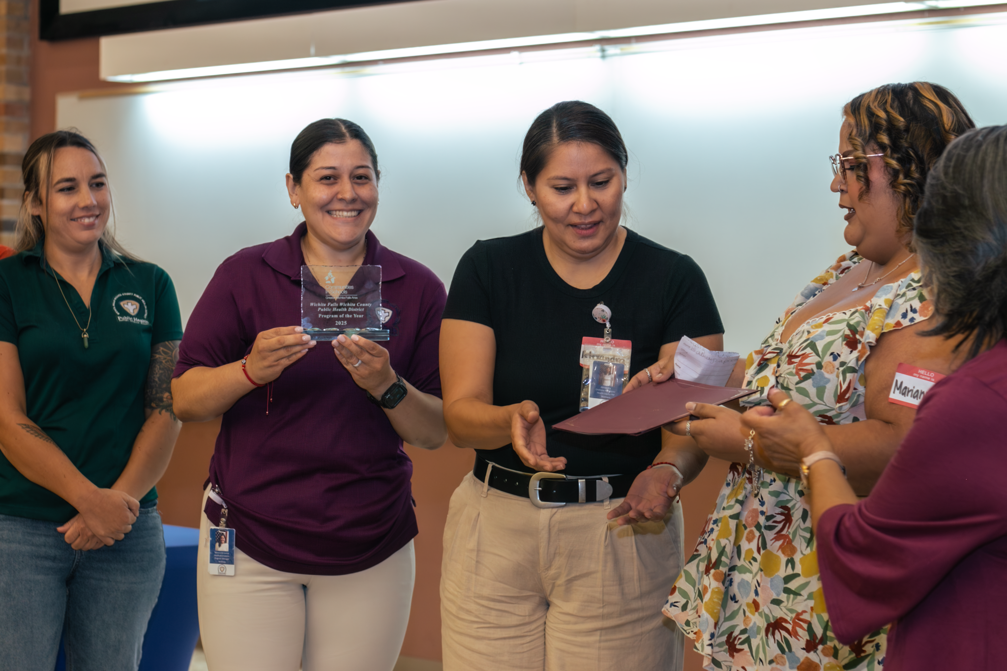 A group of women at an award ceremony, with one woman holding a glass award and others exchanging documents or certificates in an indoor setting.