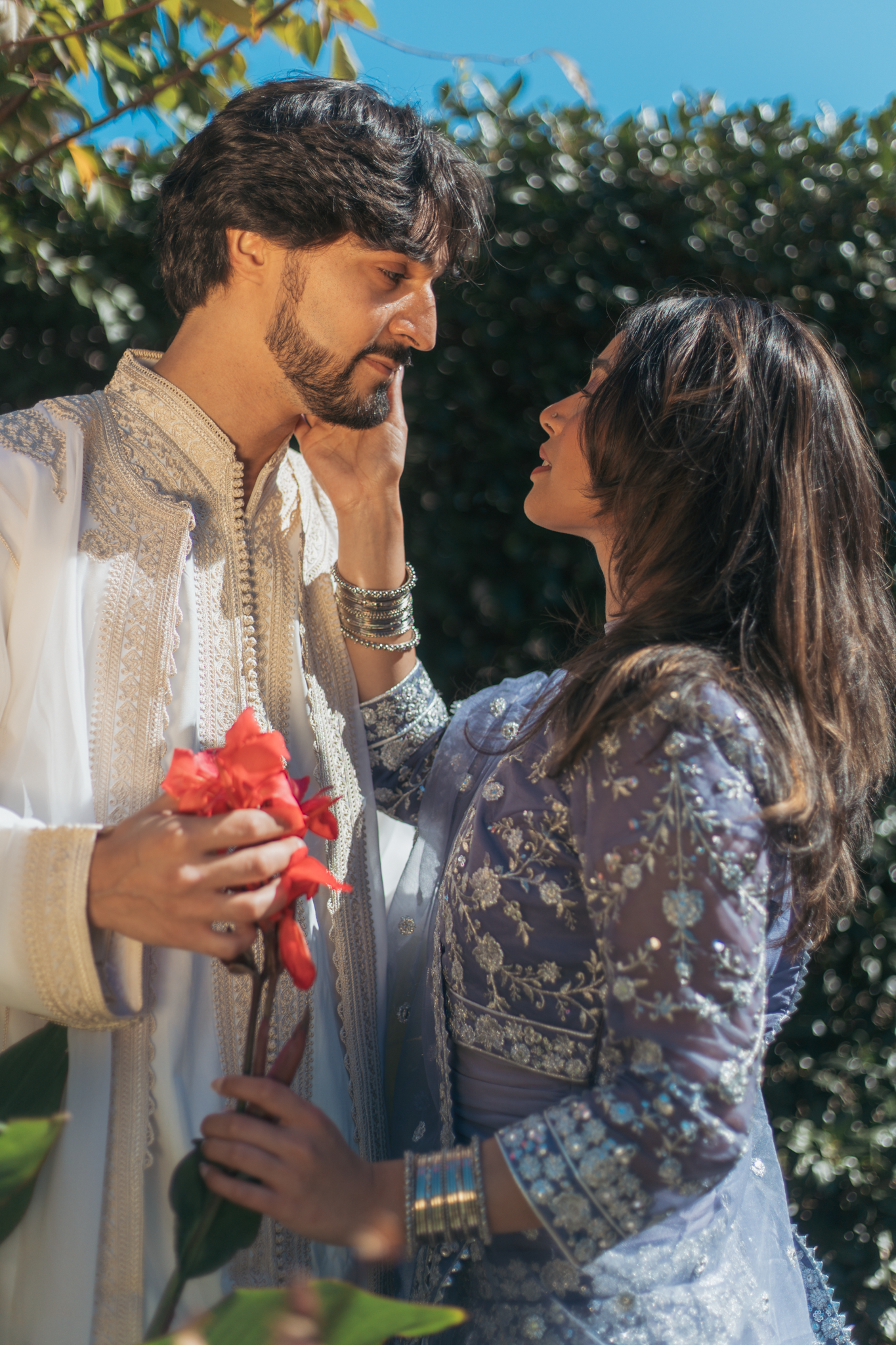 A man and woman in traditional Indian wedding attire sharing a tender moment outdoors, with the man holding pink flowers.
