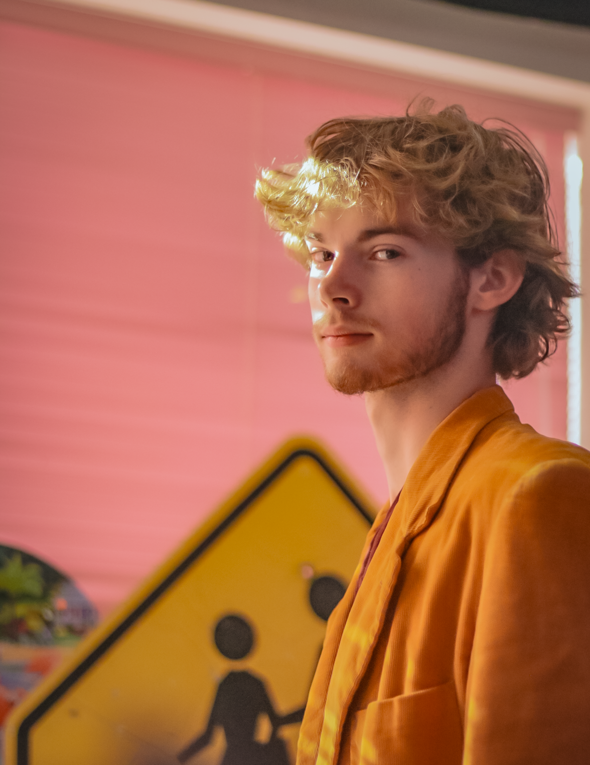 A young man with curly blonde hair and a beard looking at the camera, wearing an orange blazer, standing indoors with pink walls and a yellow pedestrian crossing sign in the background.