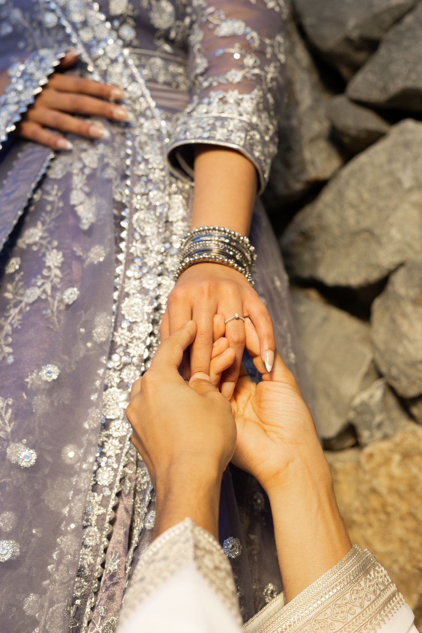 Close-up of a woman wearing a purple, heavily embellished dress with silver embroidery, as someone gently places a ring on her finger. The woman has multiple silver bangles on her wrist and her hand rests on rocks or stones.