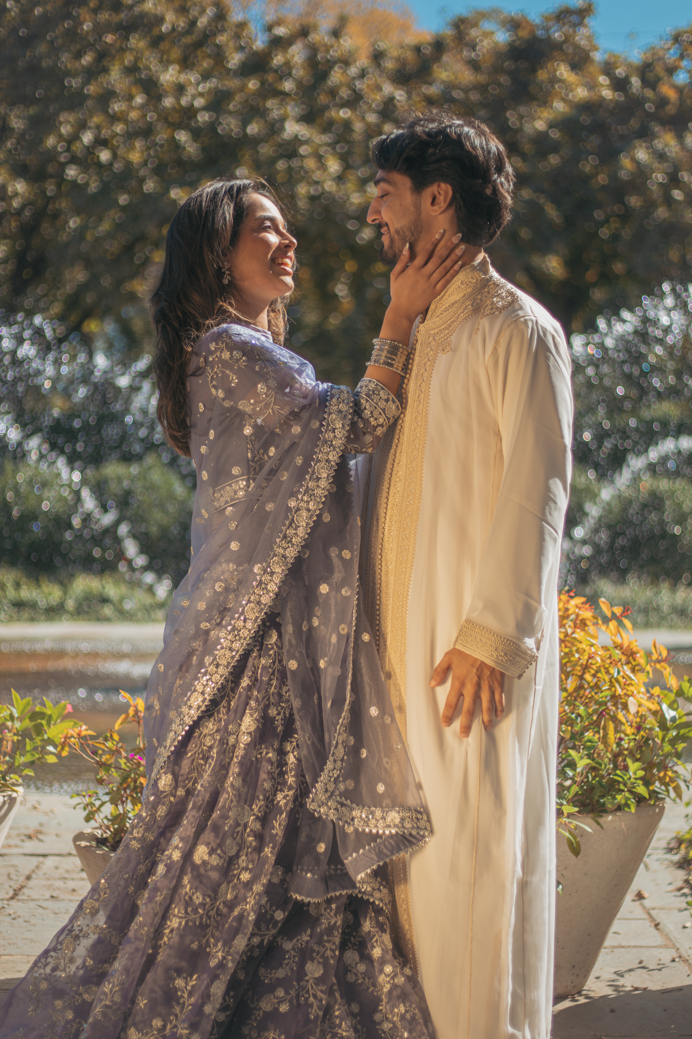 A couple wearing traditional South Asian attire standing outdoors, smiling and touching each other's faces with the woman holding the man's chin.