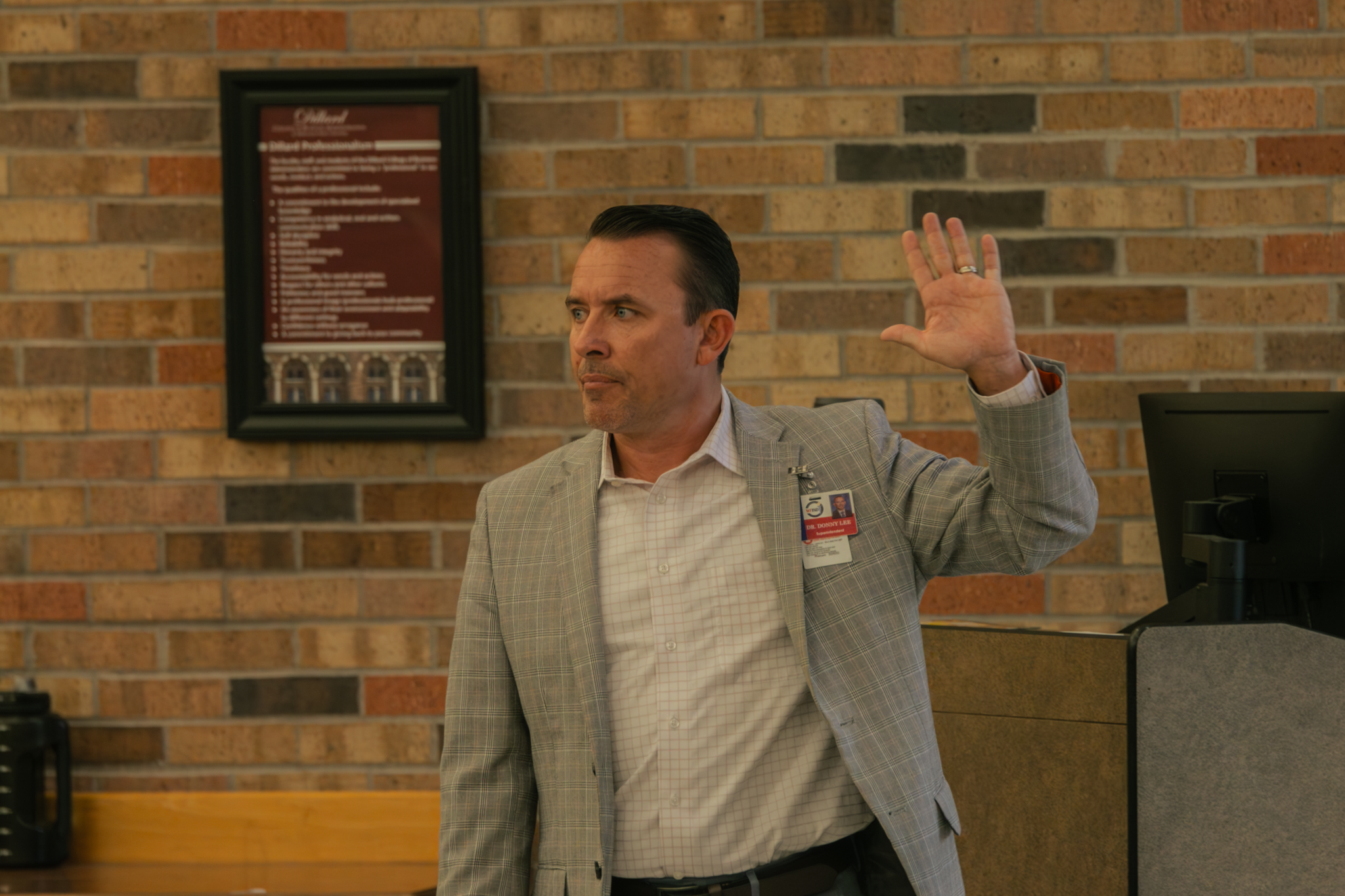 A man in a gray plaid blazer and a name badge raises his hand in a brick-walled room.