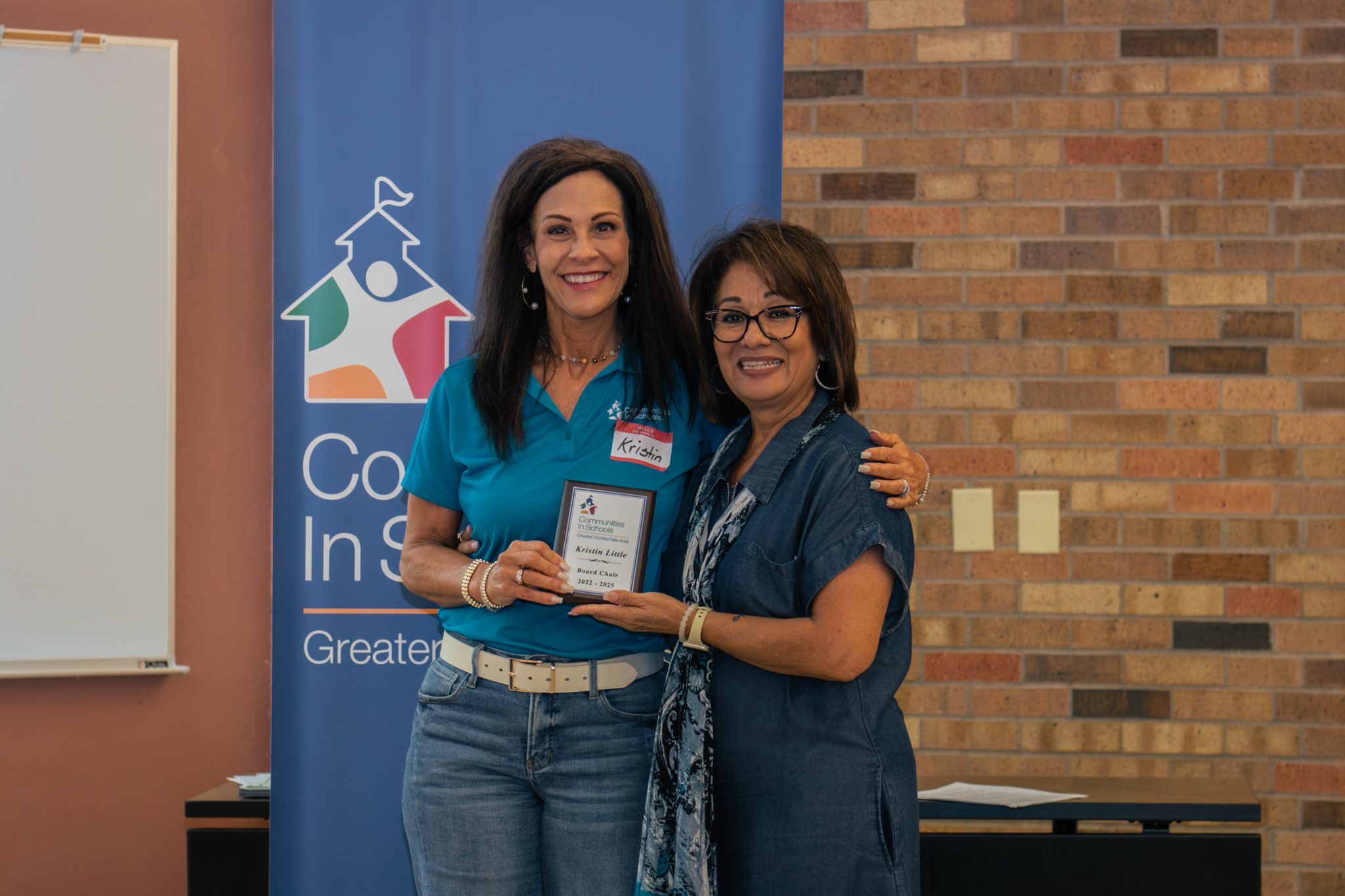 Two women smiling and holding an award plaque, standing in front of a blue banner and brick wall at an indoor event.